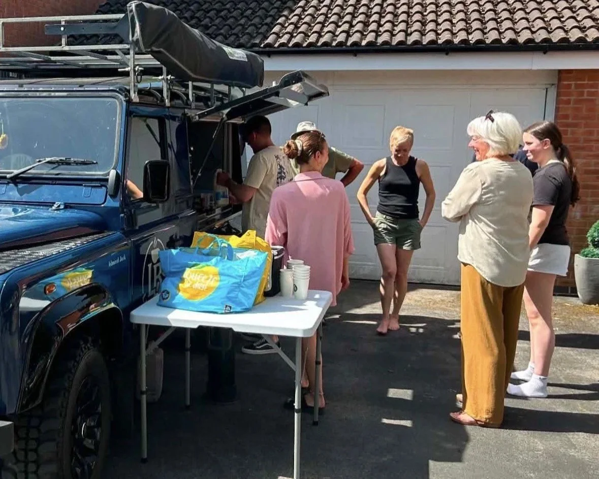 People gathered around a food truck in a driveway, with some waiting in line and others ordering. There is a table with drinks, and a garage door in the background.