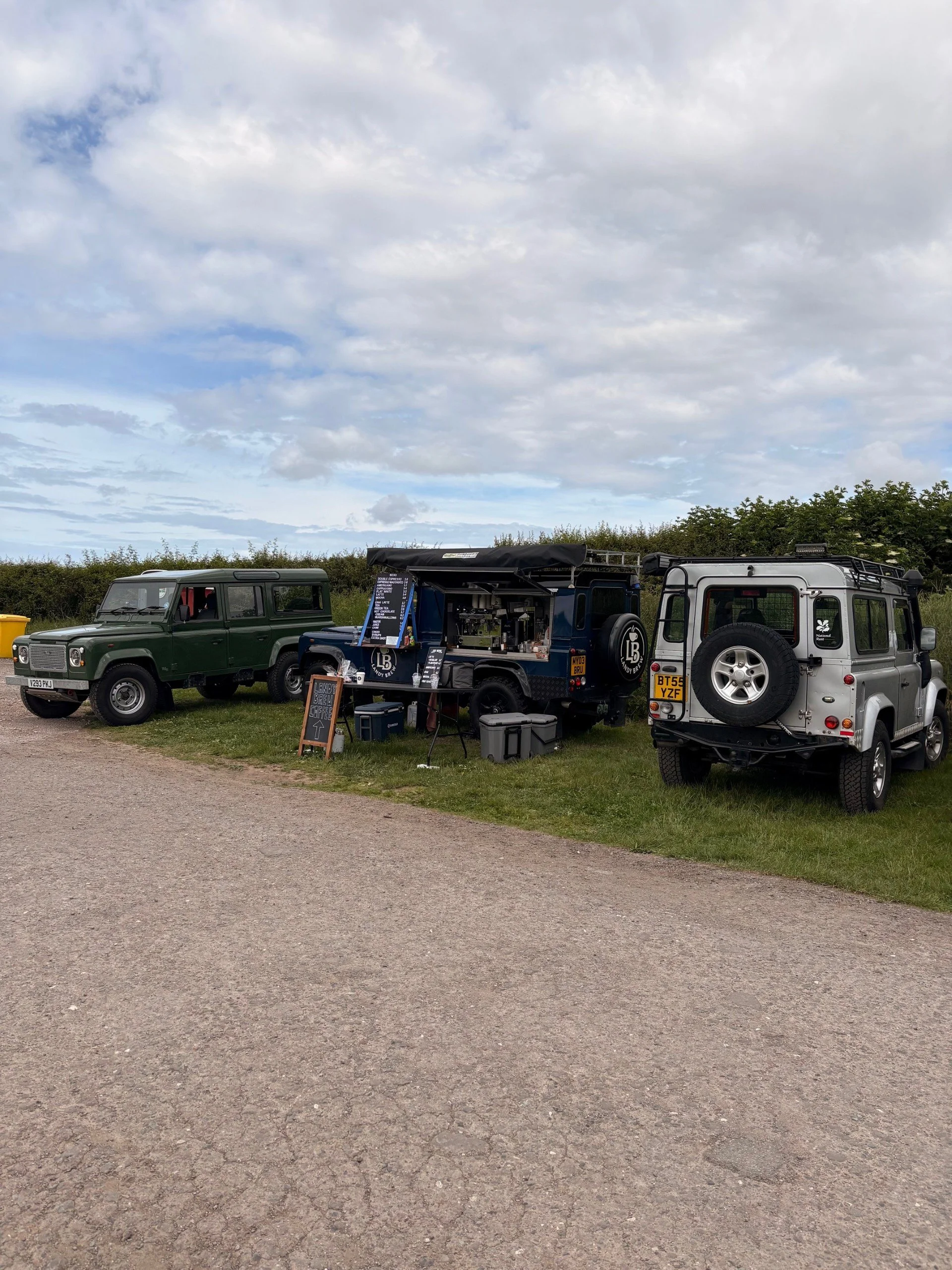 A row of three off-road vehicles parked on a grassy area under a cloudy sky. The middle vehicle is a food truck stand with a menu displayed, flanked by two other off-road vehicles, one green and one silver, with trees and bushes in the background.