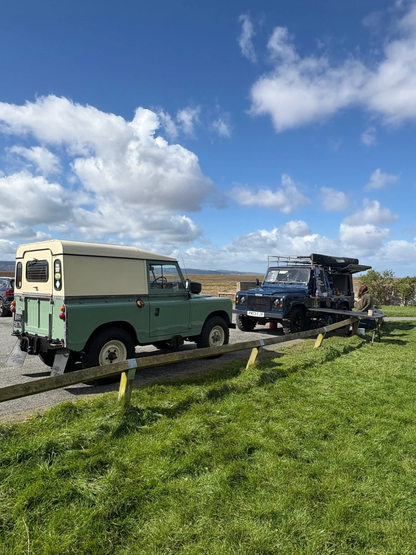 Two vintage off-road vehicles, one green and one black, parked on grass next to a wooden fence under a partly cloudy blue sky.