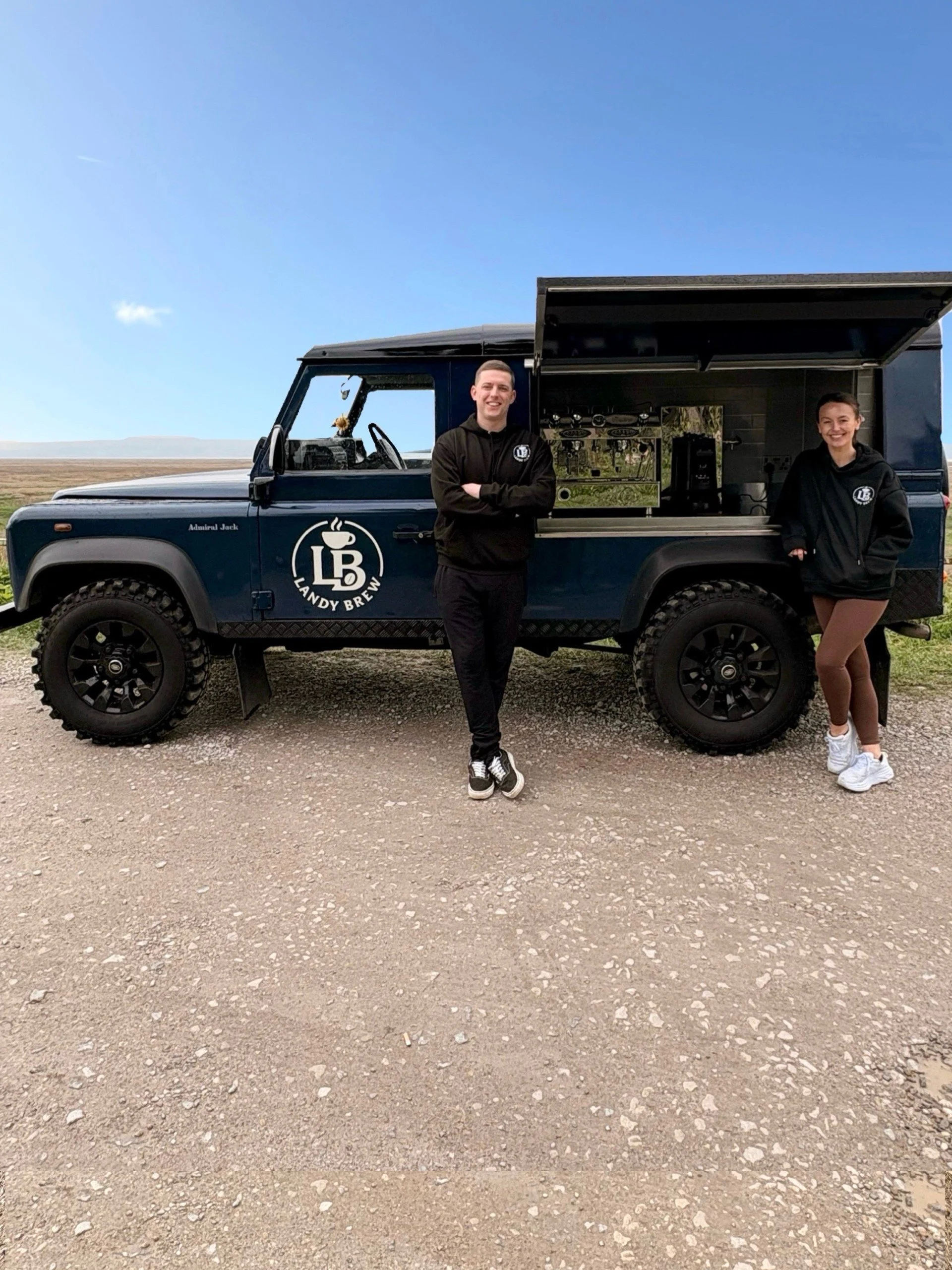 A man and a woman standing in front of a black food truck with a logo that says "Landy Brew" and a coffee cup illustration. The truck has an open side serving area. The man is smiling with arms crossed, wearing a black hoodie and pants. The woman is smiling with hands in her pockets, wearing a black hoodie, shorts, and white sneakers. The background shows a rural landscape with open fields and a blue sky.