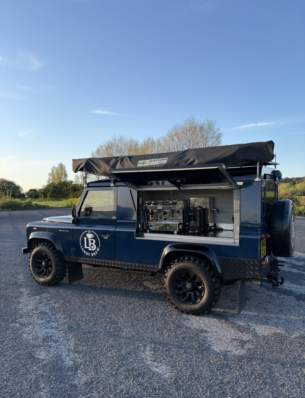 A dark blue off-road vehicle with a coffee stand setup on its side, displaying a sign that reads 'Landy Brew' with a coffee cup logo, parked on an open gravel area with trees and a clear blue sky in the background.