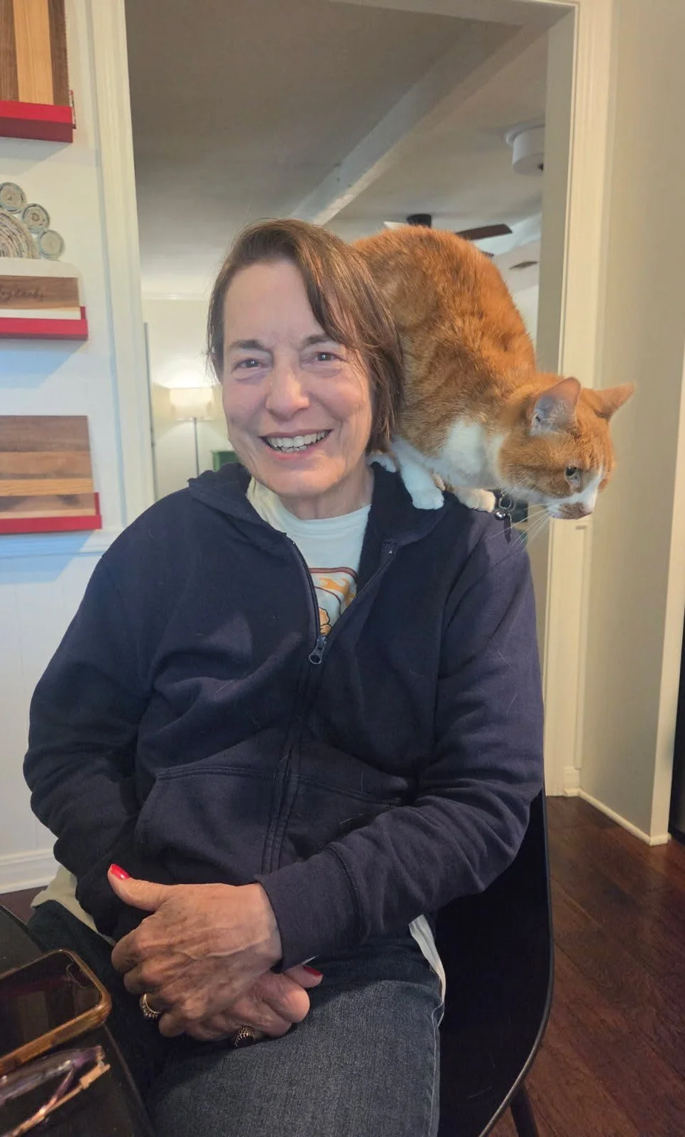 A smiling woman sitting indoors with an orange and white cat perched on her shoulder, looking to the right.