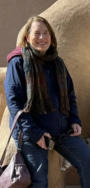 A smiling woman with shoulder-length brown hair wearing a blue jacket and multicolored scarf, sitting outdoors against a beige wall.