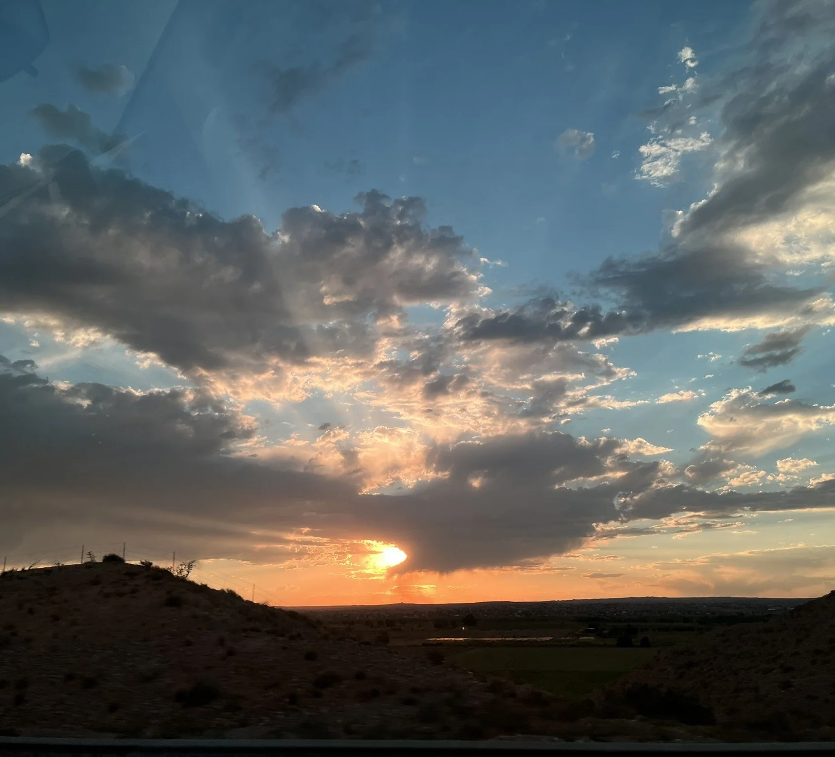 View of a sunset with partly cloudy sky over rolling hills and a distant body of water.
