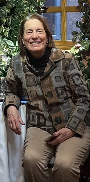 A smiling woman with short brown hair sitting in a cozy indoor setting with greenery and a wooden window in the background.