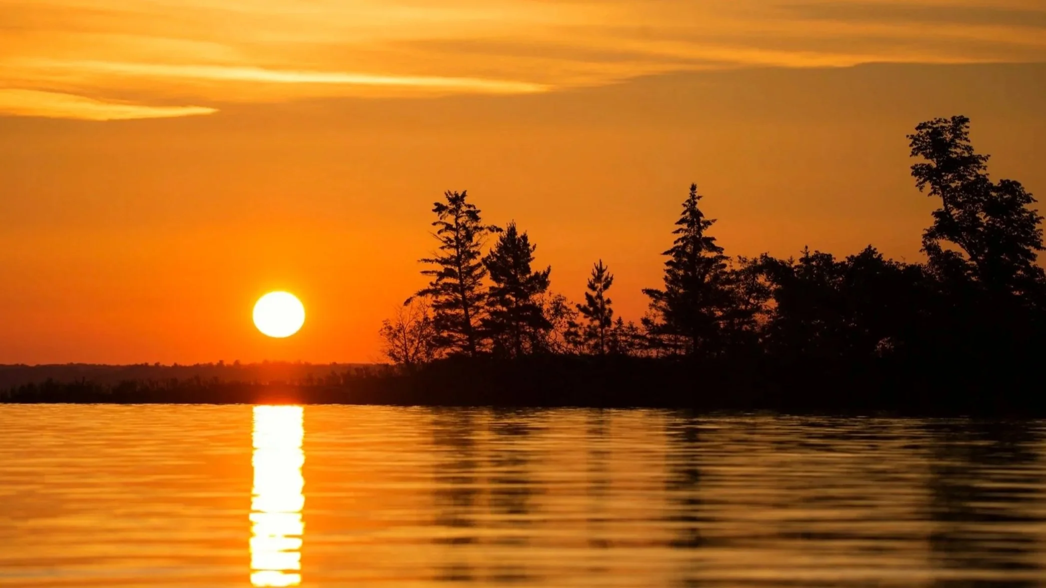 Sun setting over a body of water with a reflection, silhouetted trees on the shoreline against a colorful orange and yellow sky.
