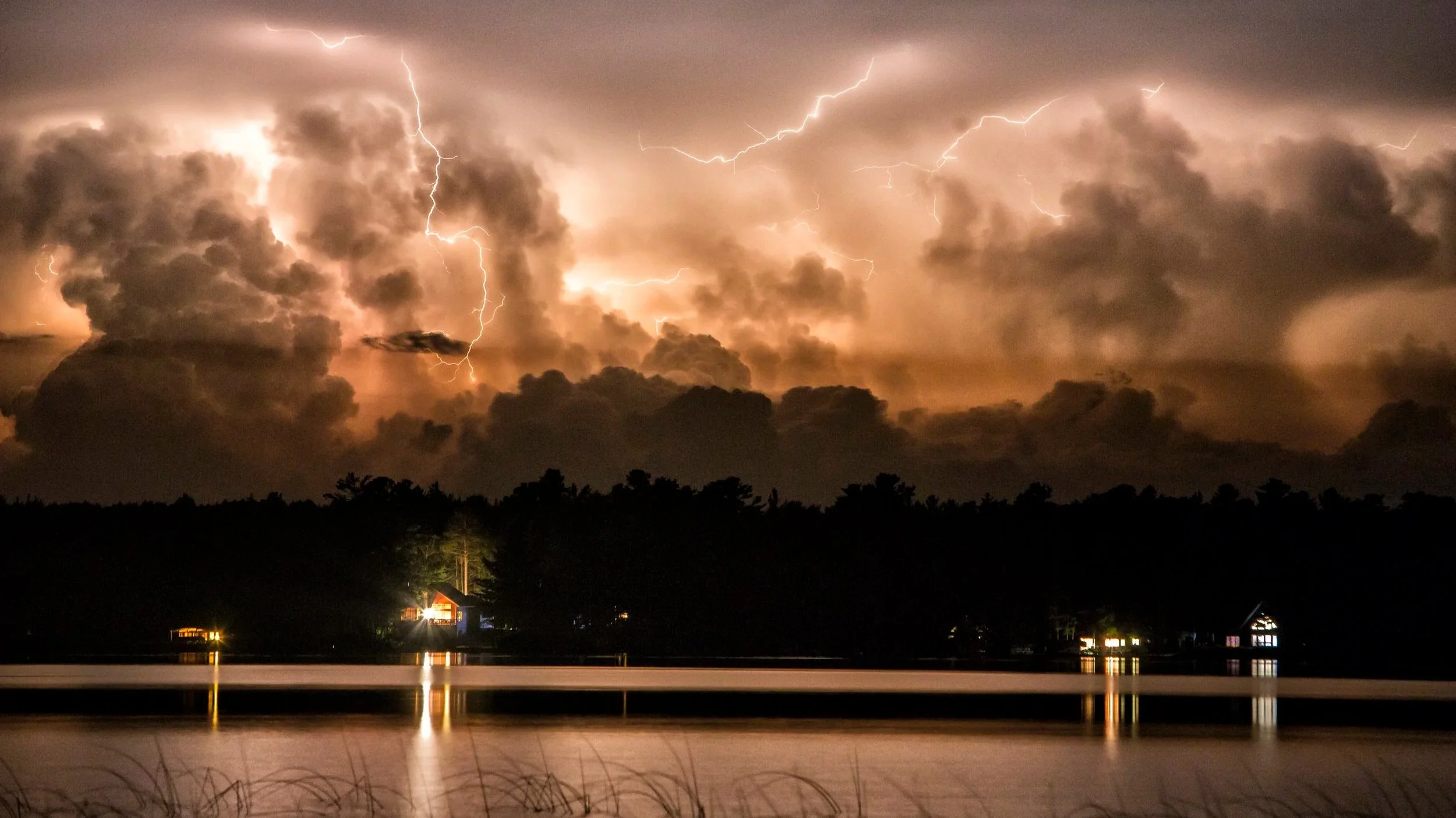 Lightning storm over a lake with reflections of houses on the water at night.