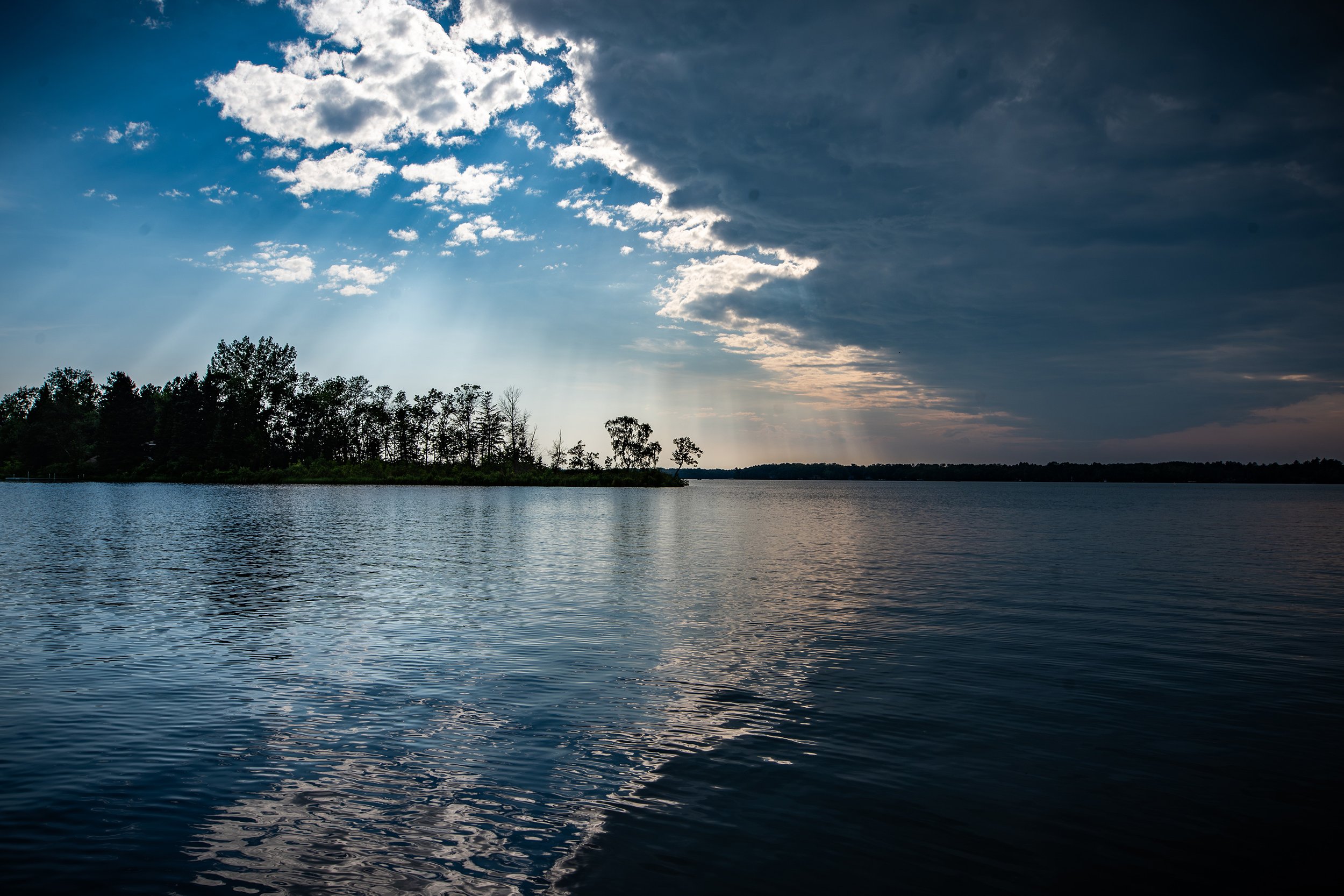 A serene lake with gentle ripples, bordered by trees and a partly cloudy sky with rays of sunlight piercing through clouds.