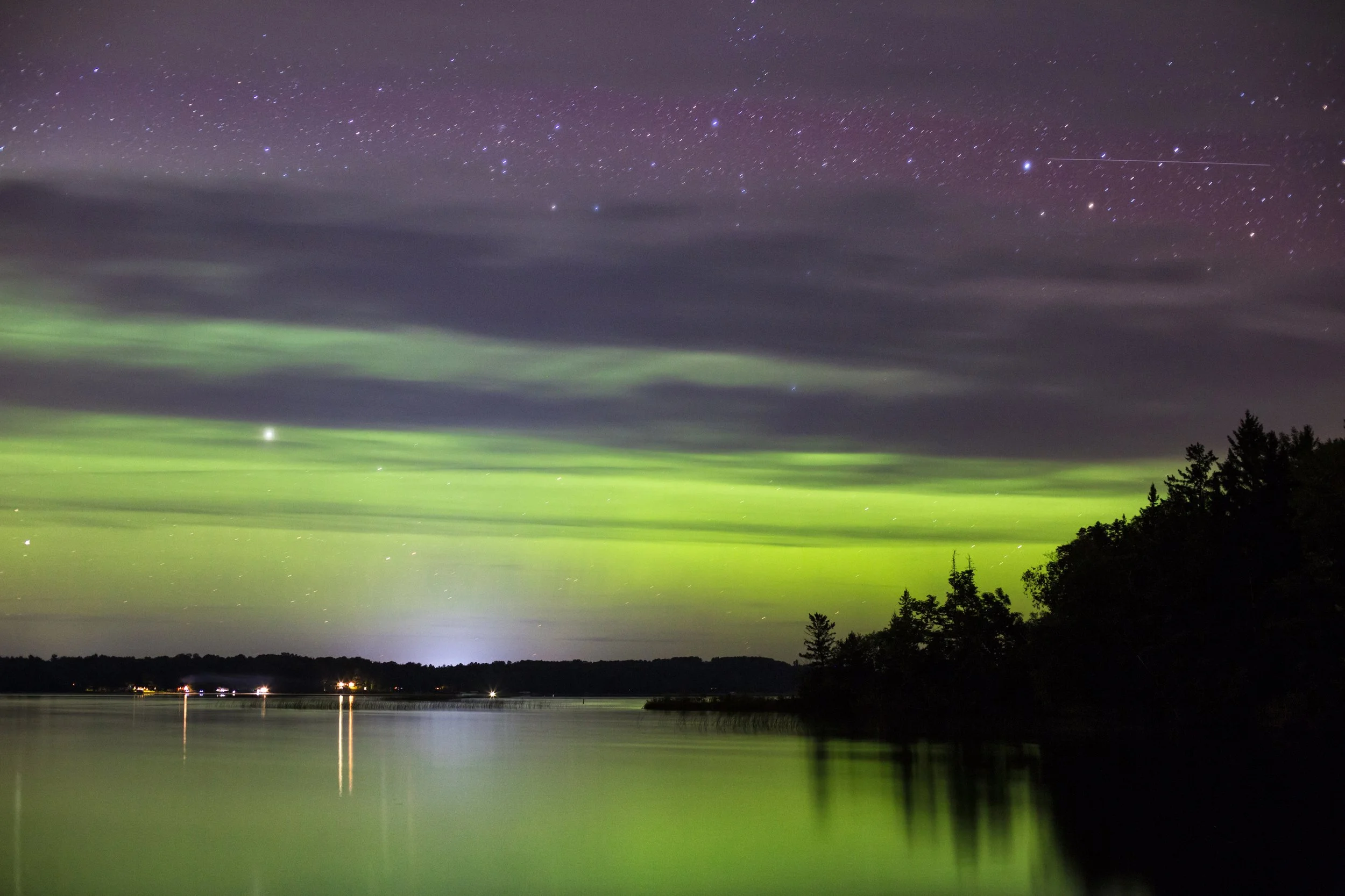 Night sky over a body of water with green aurora lights and stars, including a shooting star, and trees silhouetted at the bottom.
