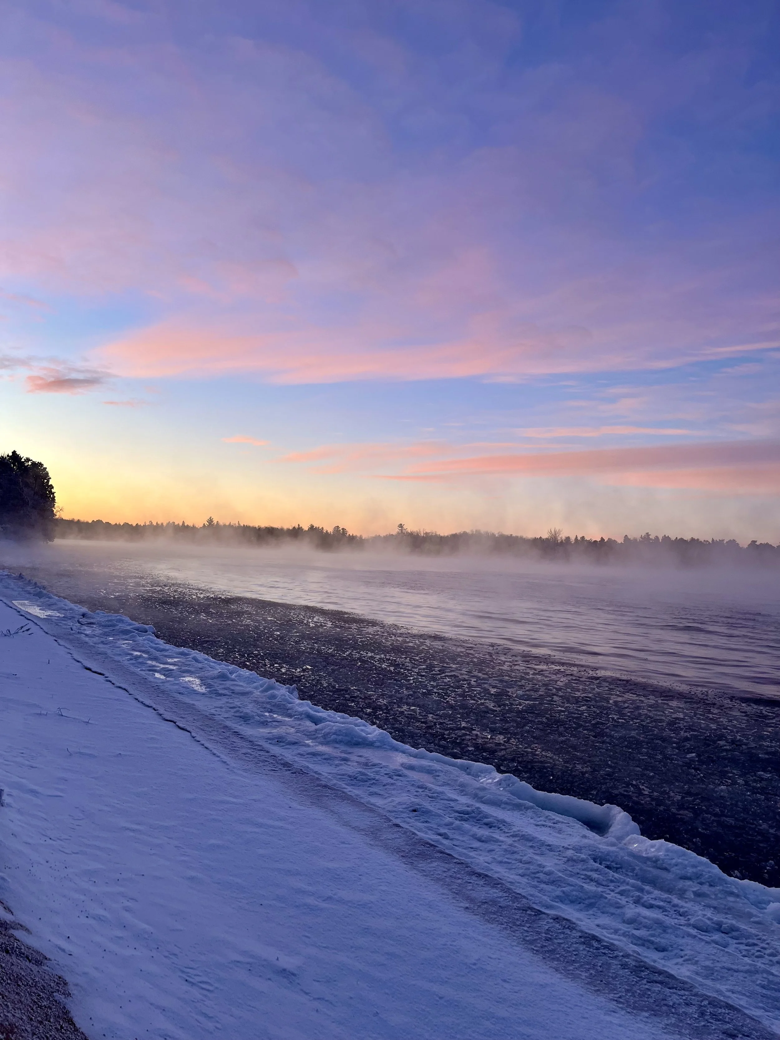 A river with mist rising above the water, surrounded by snow-covered shores and trees, under a colorful sky at dawn or dusk.