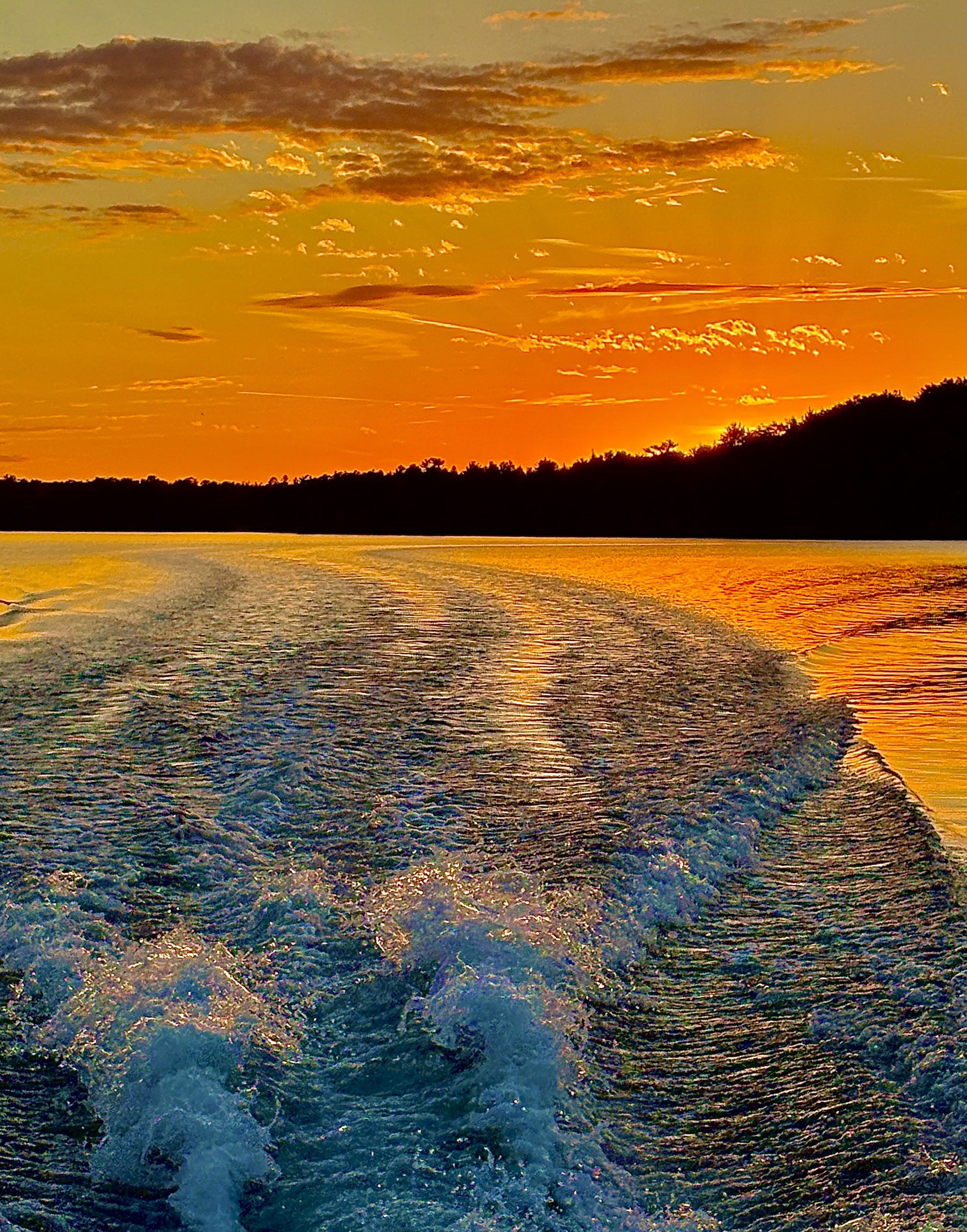 A sunset over a body of water with a silhouette of trees on the horizon and the sky filled with orange, yellow, and some dark clouds.