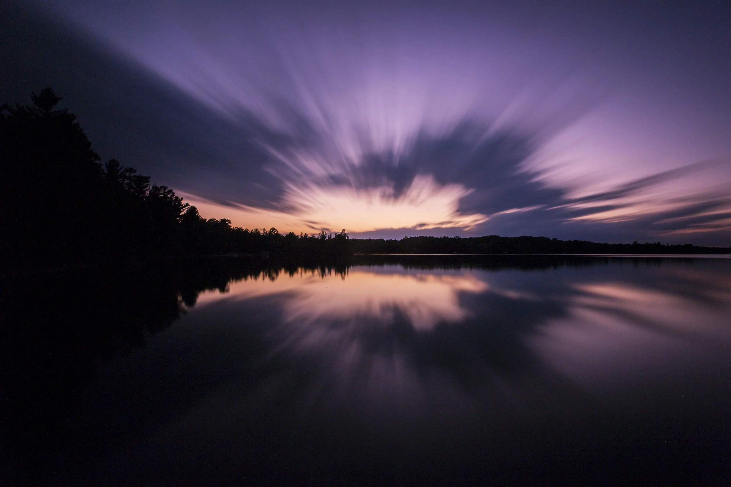 A long exposure photo of a sunset over a body of water, with the sky showing shades of purple, pink, and orange. The clouds create a streaked, dynamic pattern, and trees are silhouetted along the horizon.