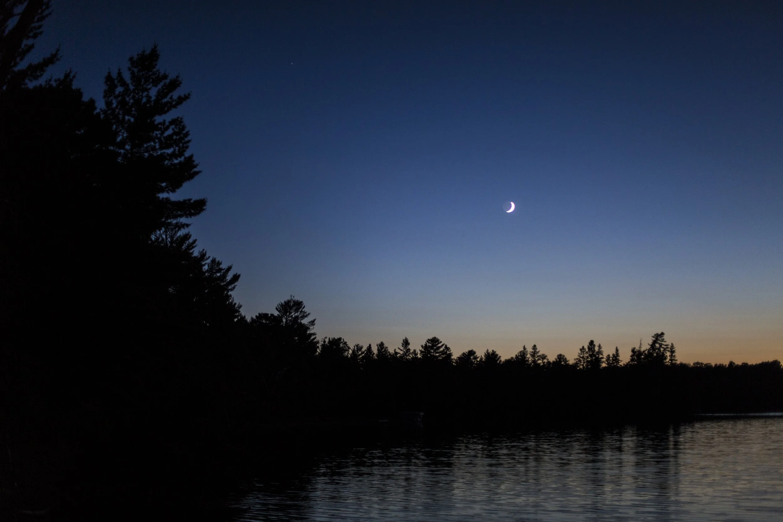 Night sky with crescent moon and a few stars, silhouette of trees along a body of water.