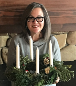 Woman with gray hair and glasses holding a Christmas wreath with four candles, seated indoors.