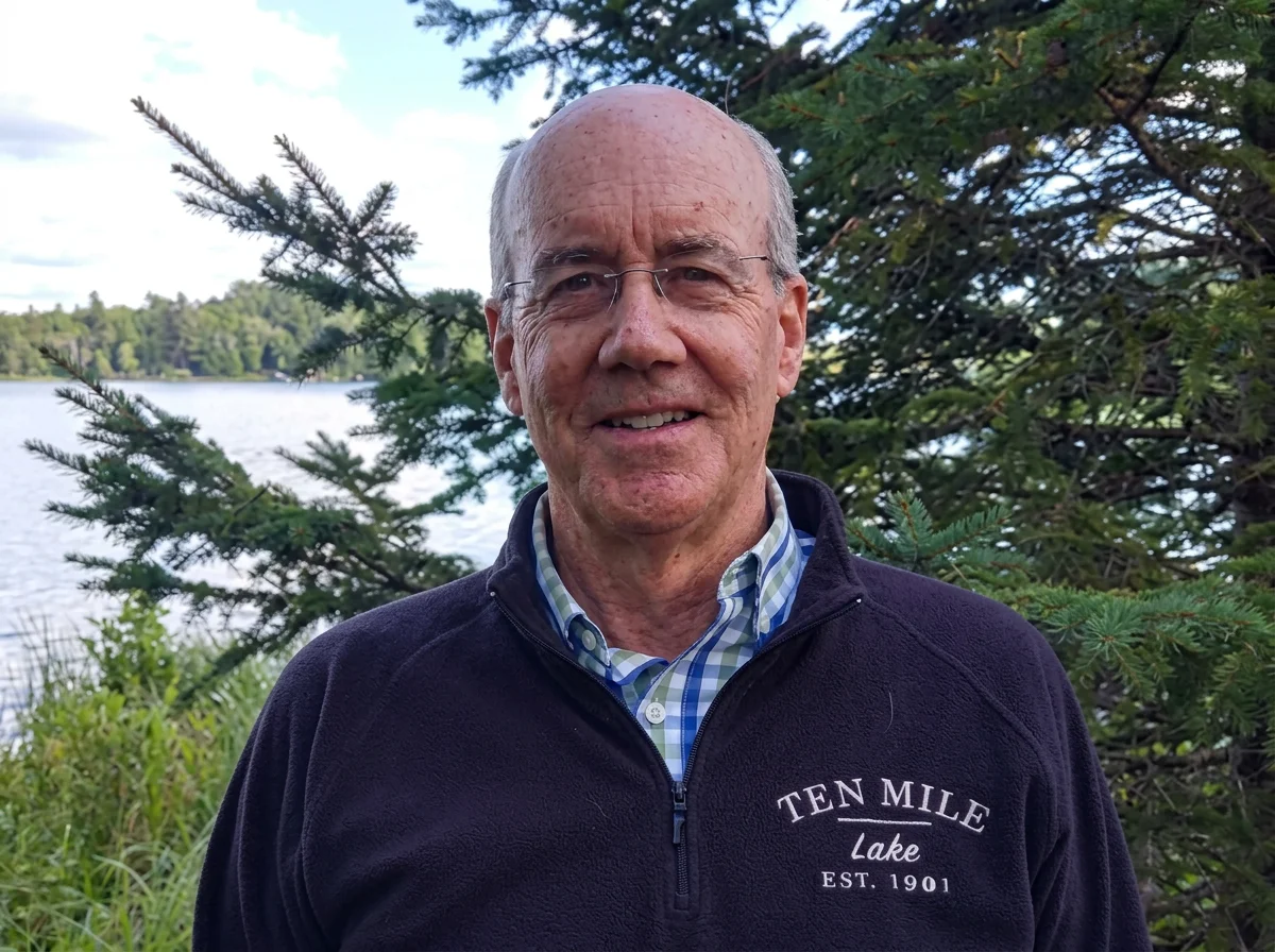 A smiling man with glasses and a receding hairline standing outdoors near a lake with trees in the background, wearing a navy quarter-zip fleece with "Ten Mile Lake Est. 1901" embroidered on it.