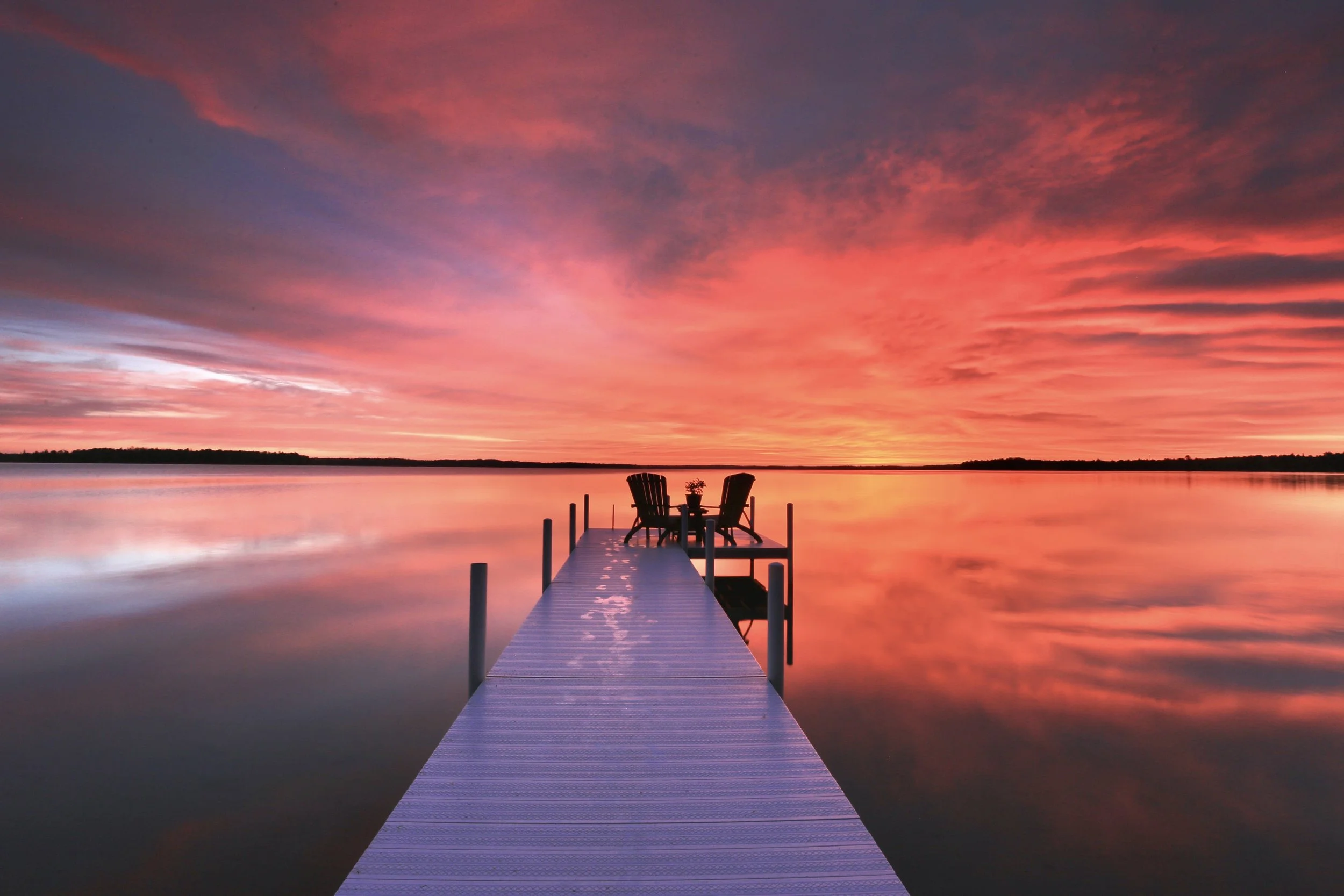 A pier extending over a calm body of water during sunset, with two chairs and a small table at the end, under a vibrant sky with pink, purple, and orange clouds.