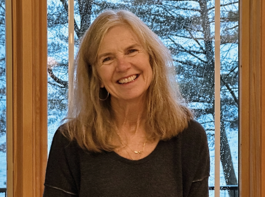 A smiling older woman with blonde hair standing indoors near a window with rain droplets, trees visible outside.