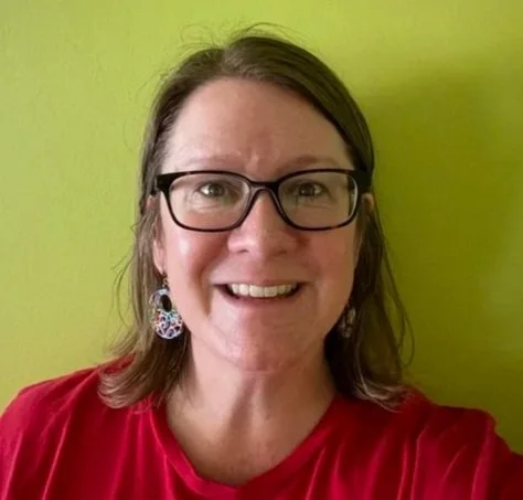 A woman with glasses, earrings, and a red shirt smiling in front of a green wall.