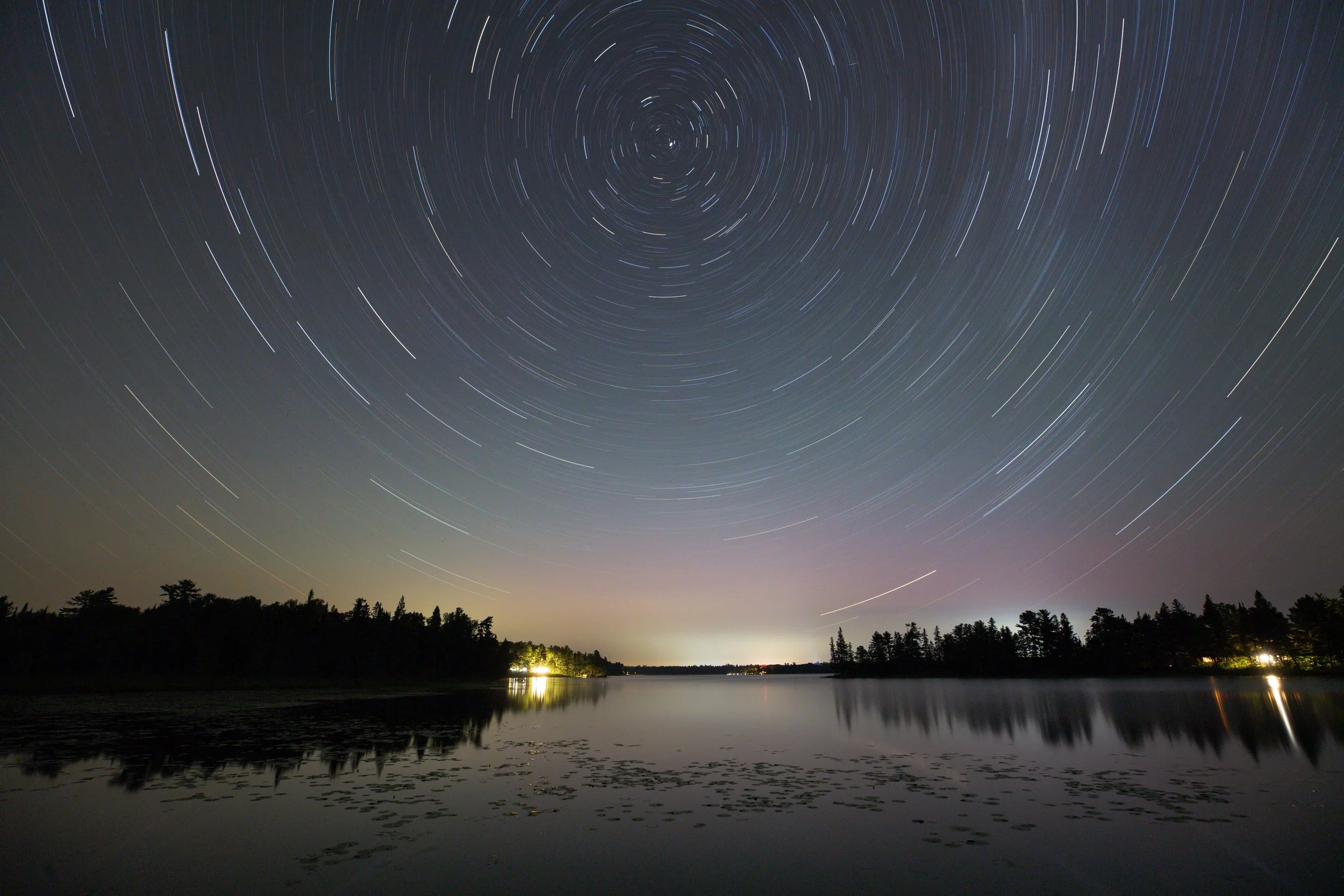 Long exposure photograph of star trails in the night sky over a lake, with trees silhouetted along the water's edge and a faint glow on the horizon.