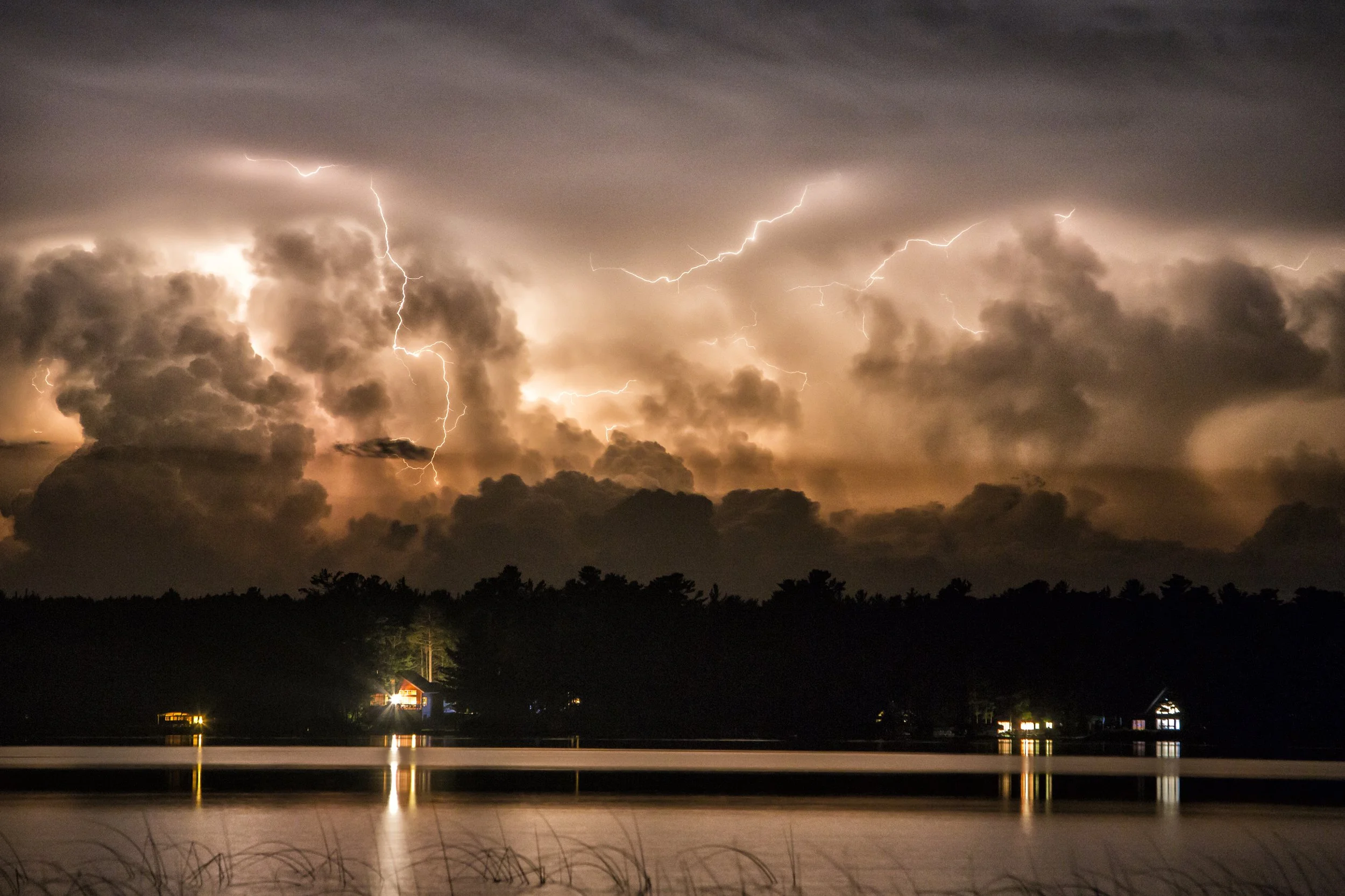 Lightning storm over a lake with a dark cloudy sky, silhouettes of trees and houses with reflections on water.