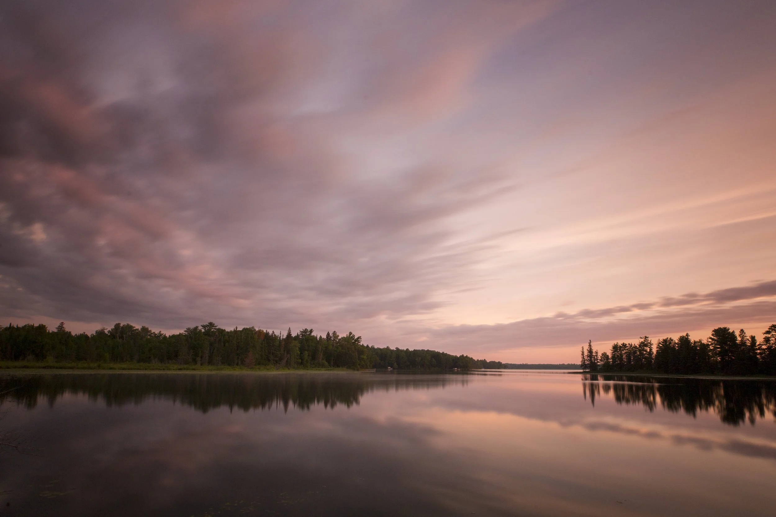 Sunset over a lake with a forested shoreline and pink, purple, and orange clouds in the sky.