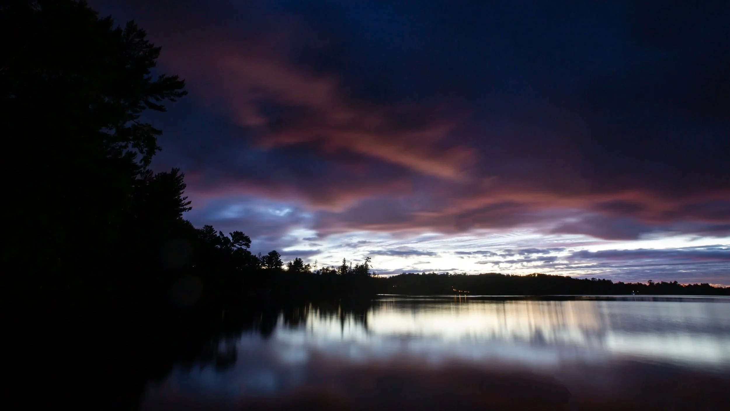 A serene landscape with a river reflecting a colorful sky during sunset, with dark silhouettes of trees on the left.