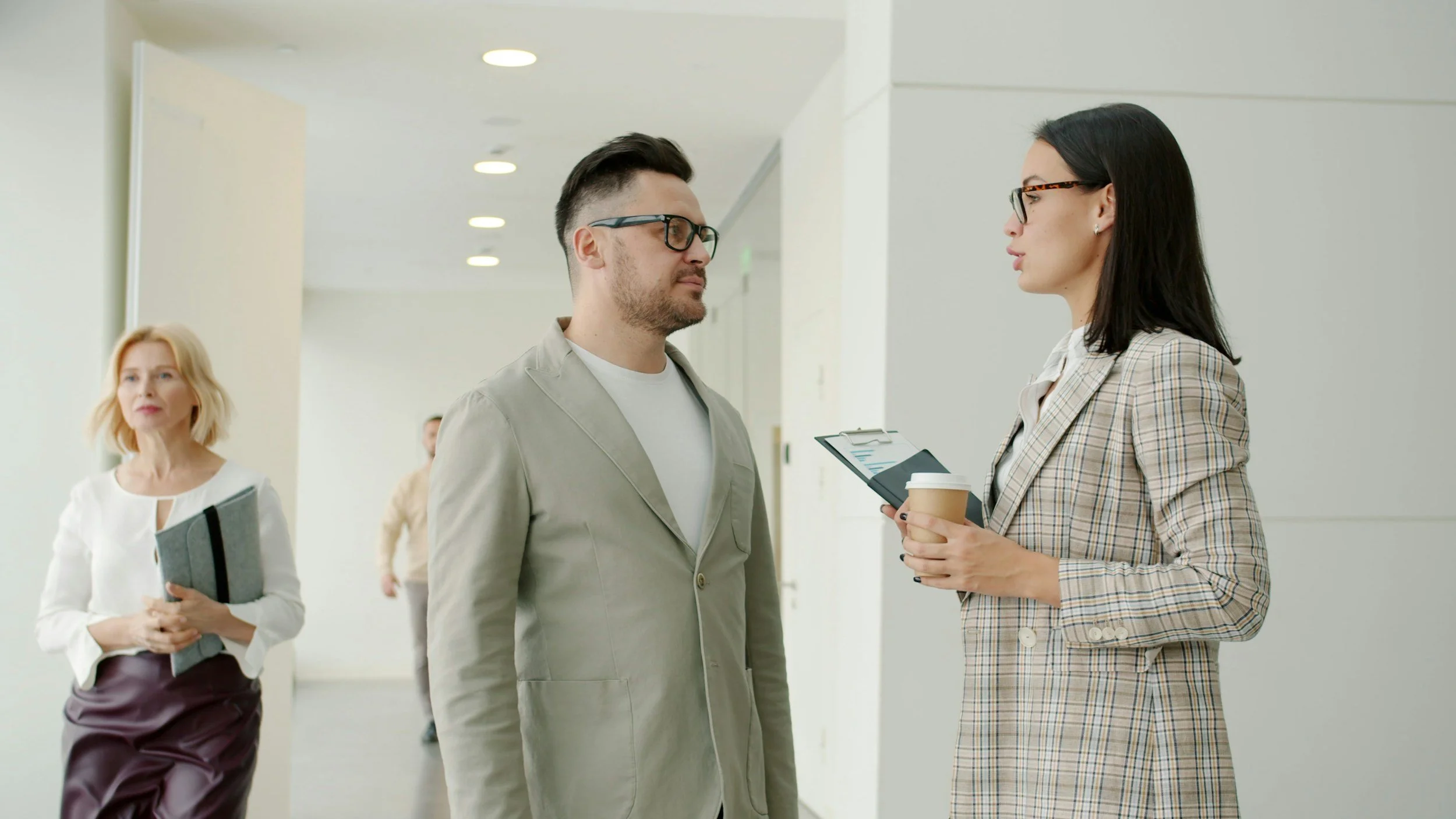 Two professionals, a man and a woman, having a conversation in an office hallway, while a third woman stands nearby holding a folder, during a break or meeting.