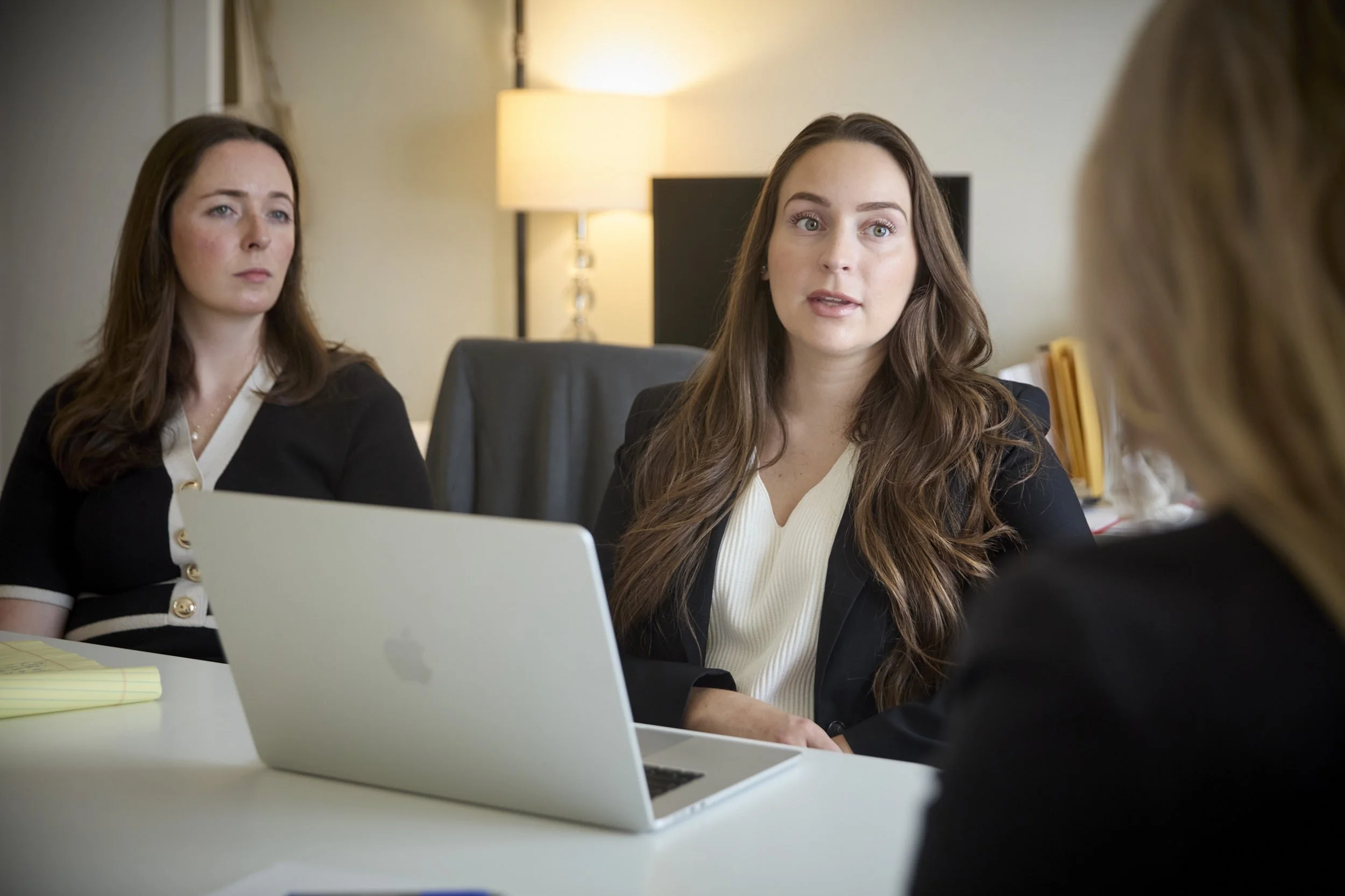Three women sitting at a table in an office, engaged in conversation. One woman with long, wavy brown hair and wearing a black blazer with a white top is speaking, while another woman with straight brown hair and a black cardigan looks on. A third woman with blonde hair is partially visible in the foreground, facing away from the camera. There is a laptop, yellow notepad, and lamp in the background.