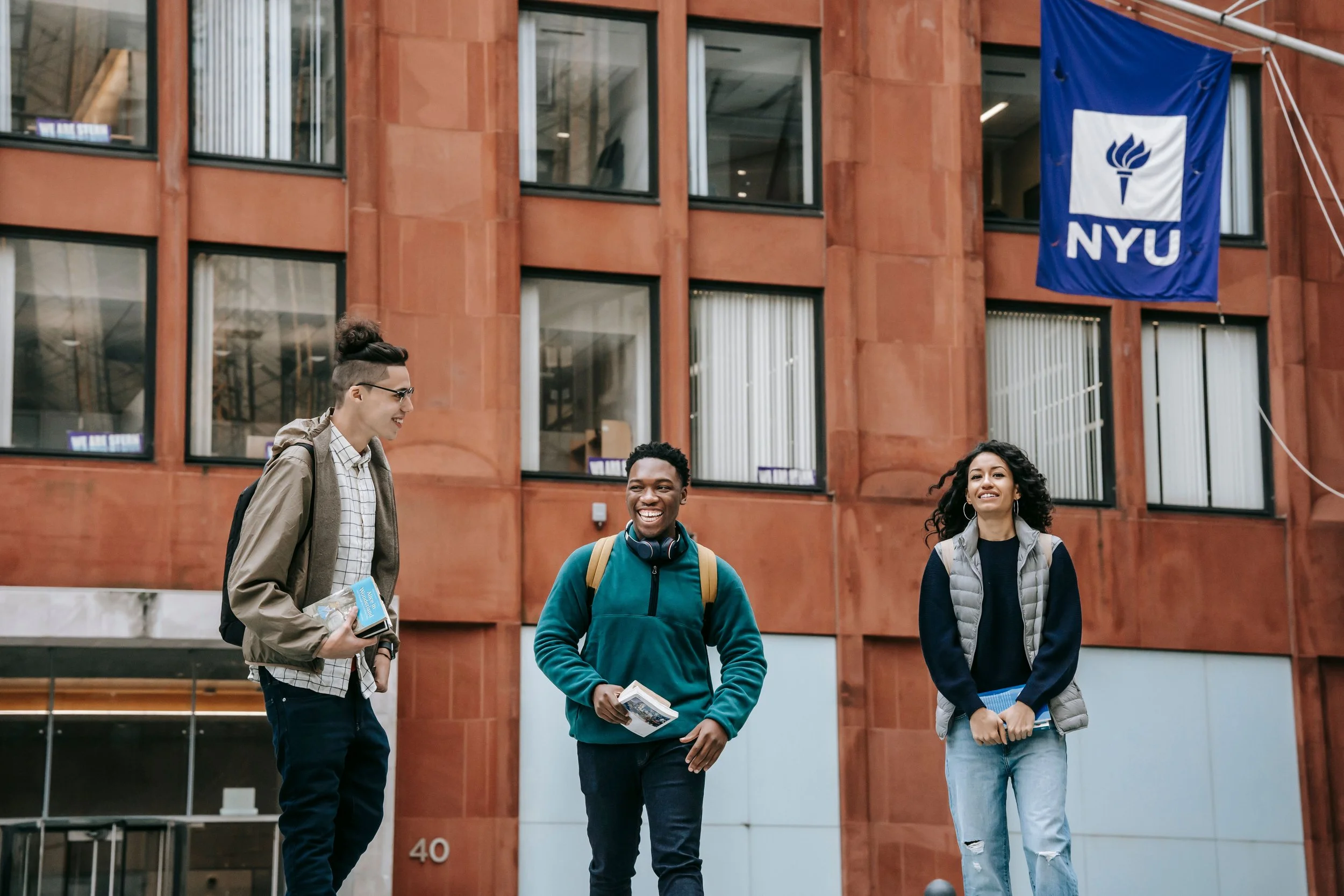 Three ethnically diverse students smile and walk together outside a building with a NYU banner, carrying books and notebooks.