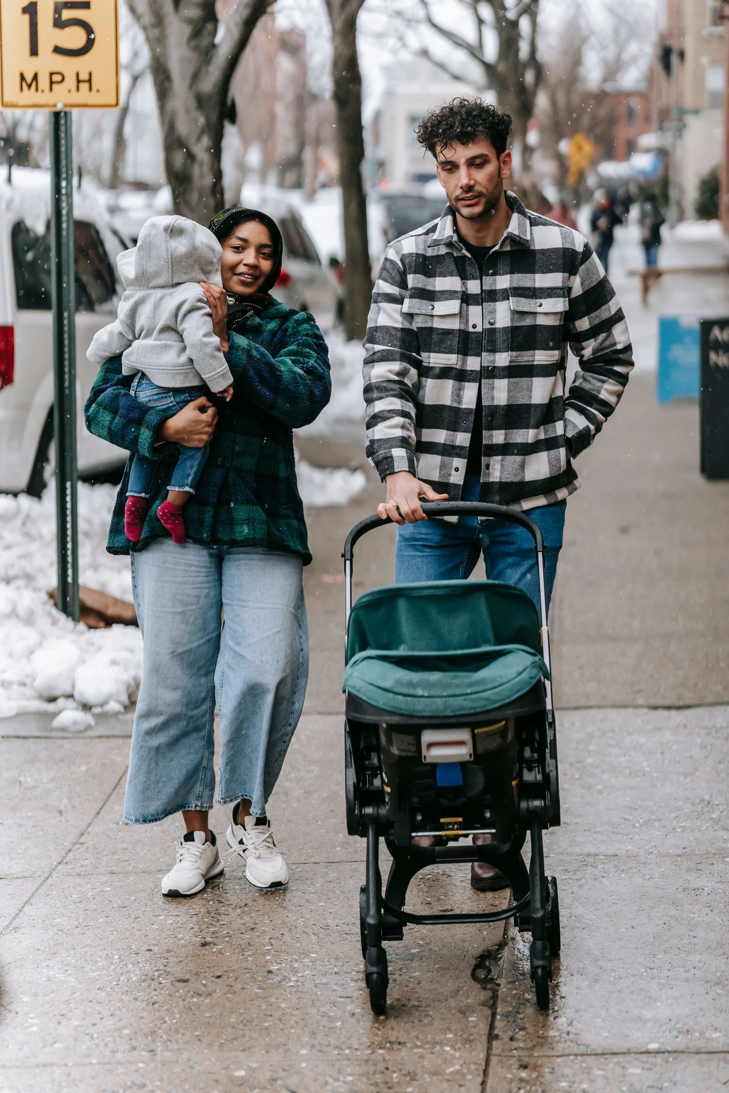 A light-brown skinned family walk towards the camera on a snow-dusted sidewalk. The woman is carrying a child and smiling, while the man is pushing a stroller. The scene is outdoors on a city street with parked cars and leafless trees.