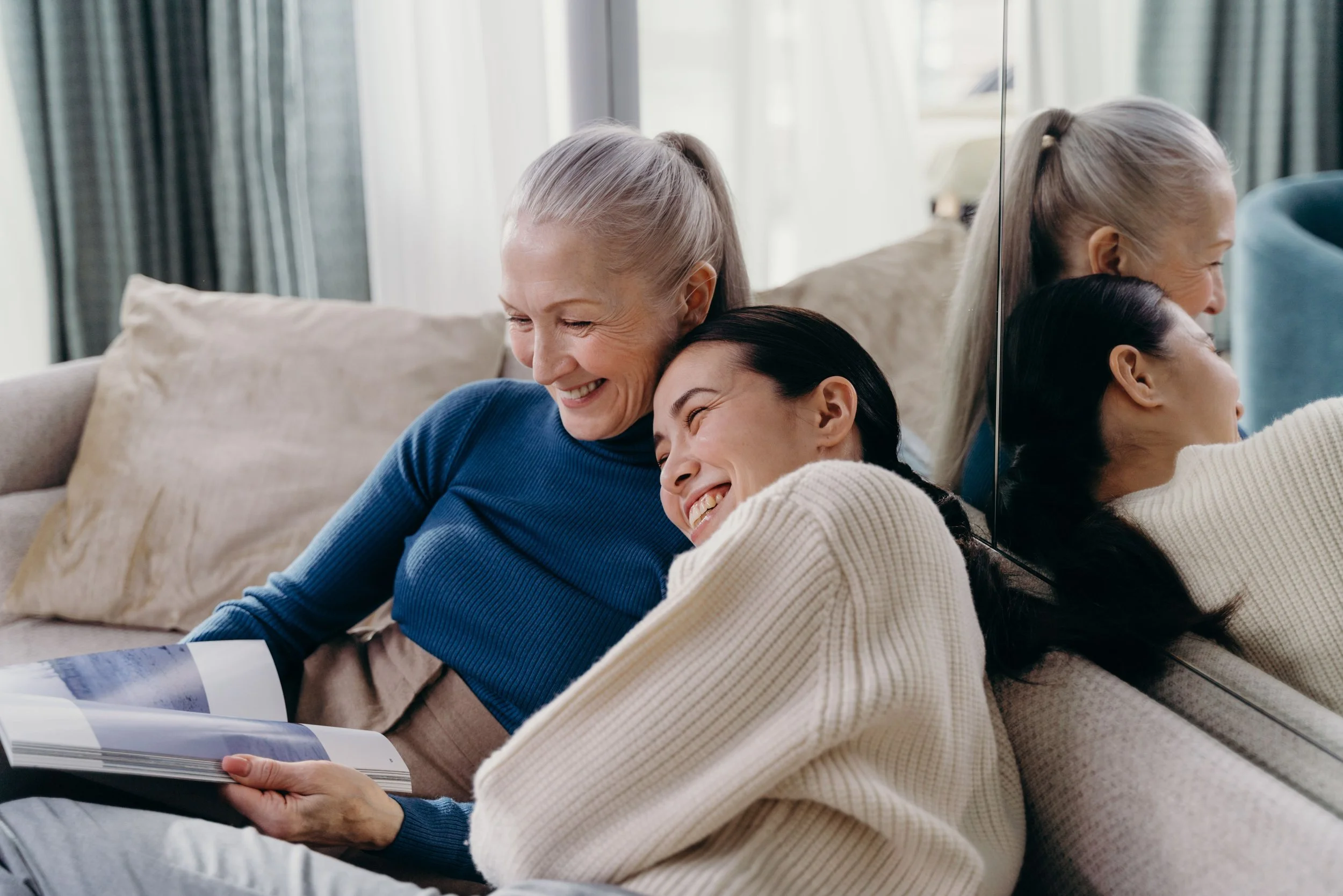 Three women sitting on a beige couch, smiling and sharing a moment of joy. One woman with gray hair and wearing a blue sweater is hugging another woman with dark hair wearing a white sweater. A third woman with gray hair, also with a ponytail, is leaning on the couch and looking at the book the second woman is holding.