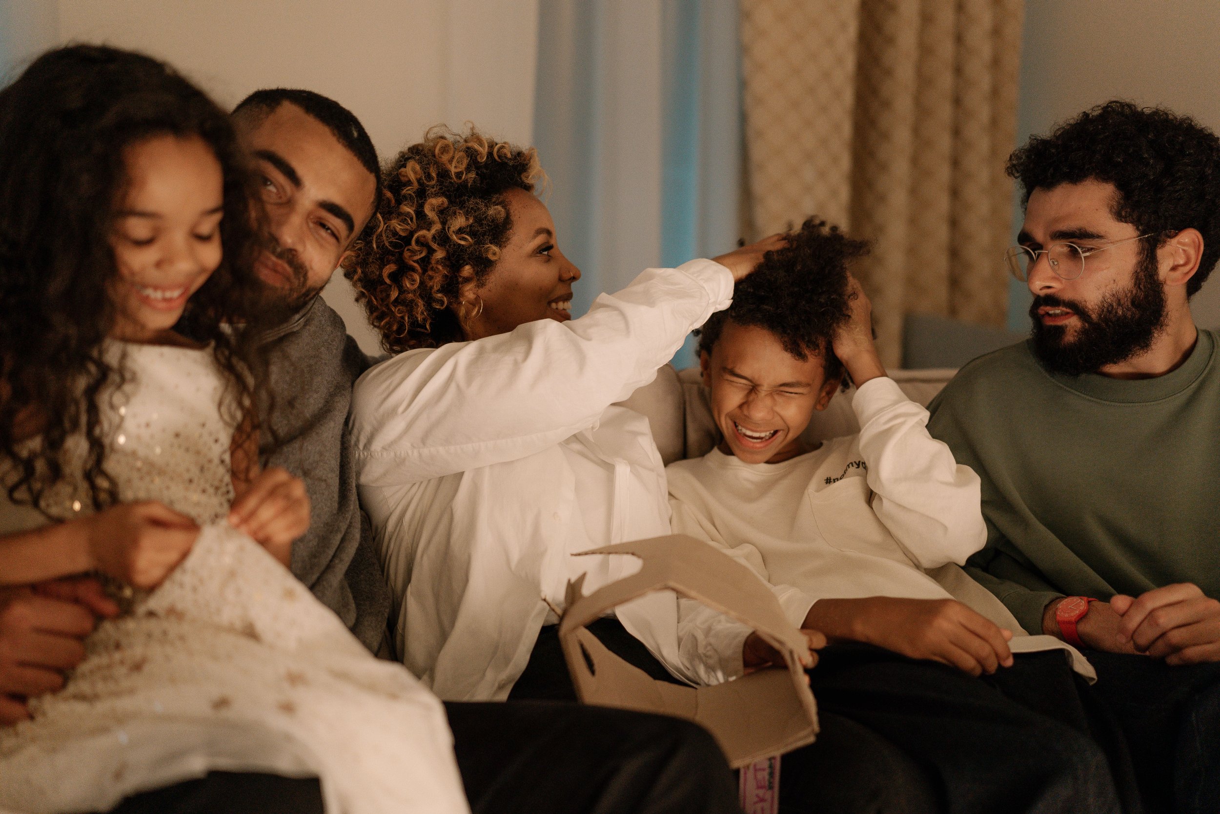 A Black family, sitting closely together on a couch, smiling and joking around, including two young children, a young adult, a mother and a father, with a warm, cozy indoor setting in the background.