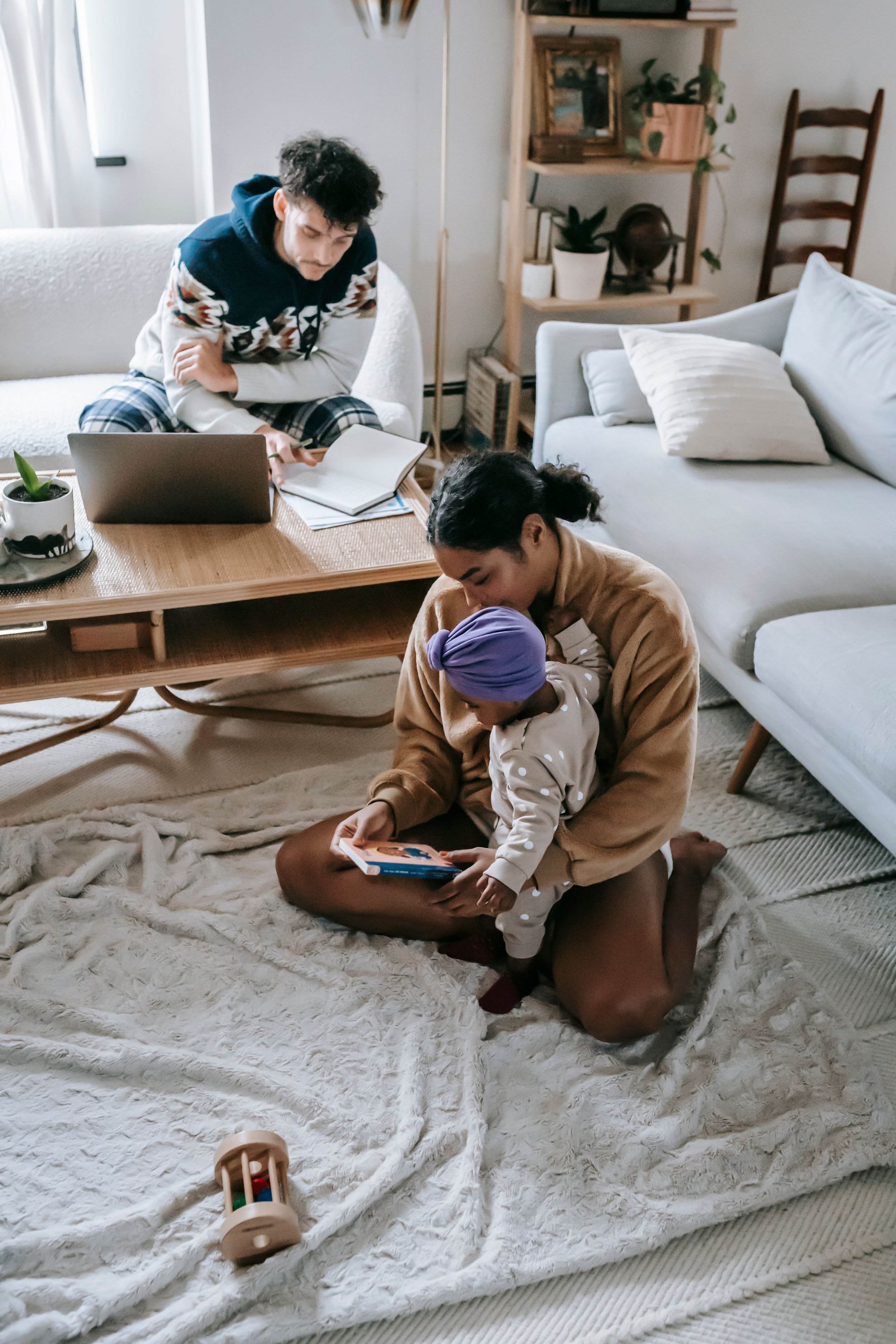 A Black mother sits on the floor with her child on her lap, reading a book together. In the background, the White father is sitting on a sofa behind a low table, looking down at papers. The room has a cozy, modern decor.