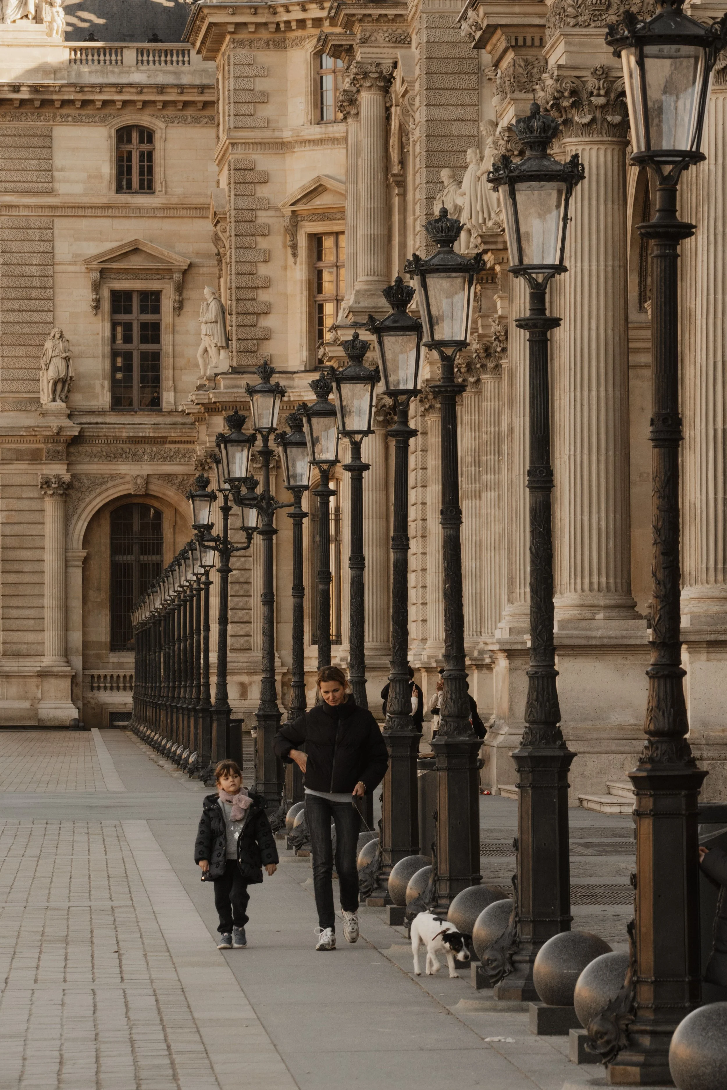 A woman and a child walking along a sidewalk in front of a historic building with ornate architectural details, sculptures, and tall black lampposts. The woman is dressed in dark clothing and the girl is wearing a black coat with a hood.