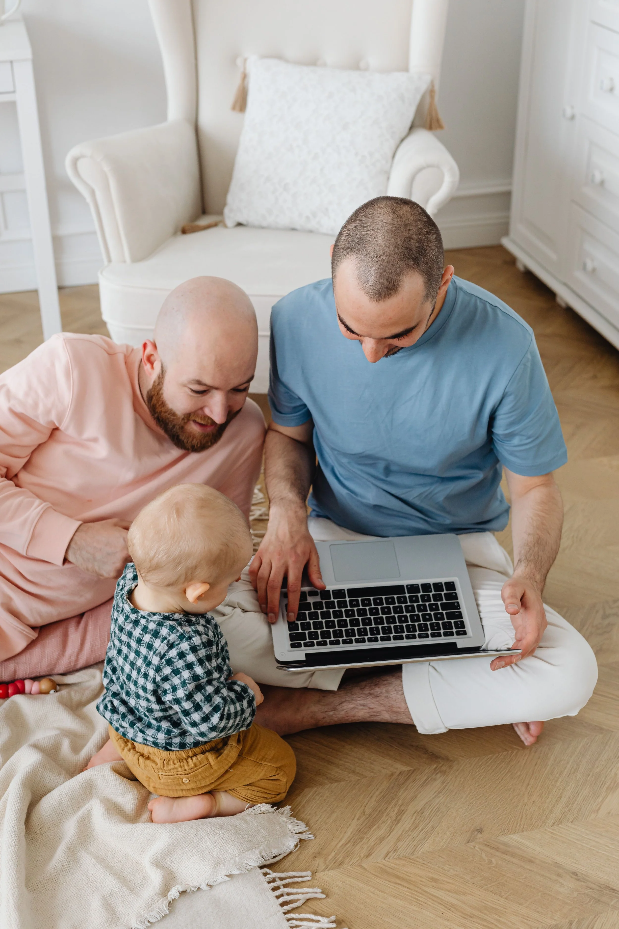 Two adult white men and their young white child sitting on the floor looking at a laptop, with a white armchair and dresser in the background.