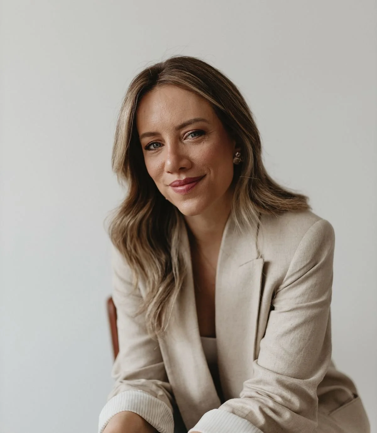 A woman with shoulder-length wavy hair, wearing a beige blazer and a white top, sitting and smiling softly against a plain light-colored background.