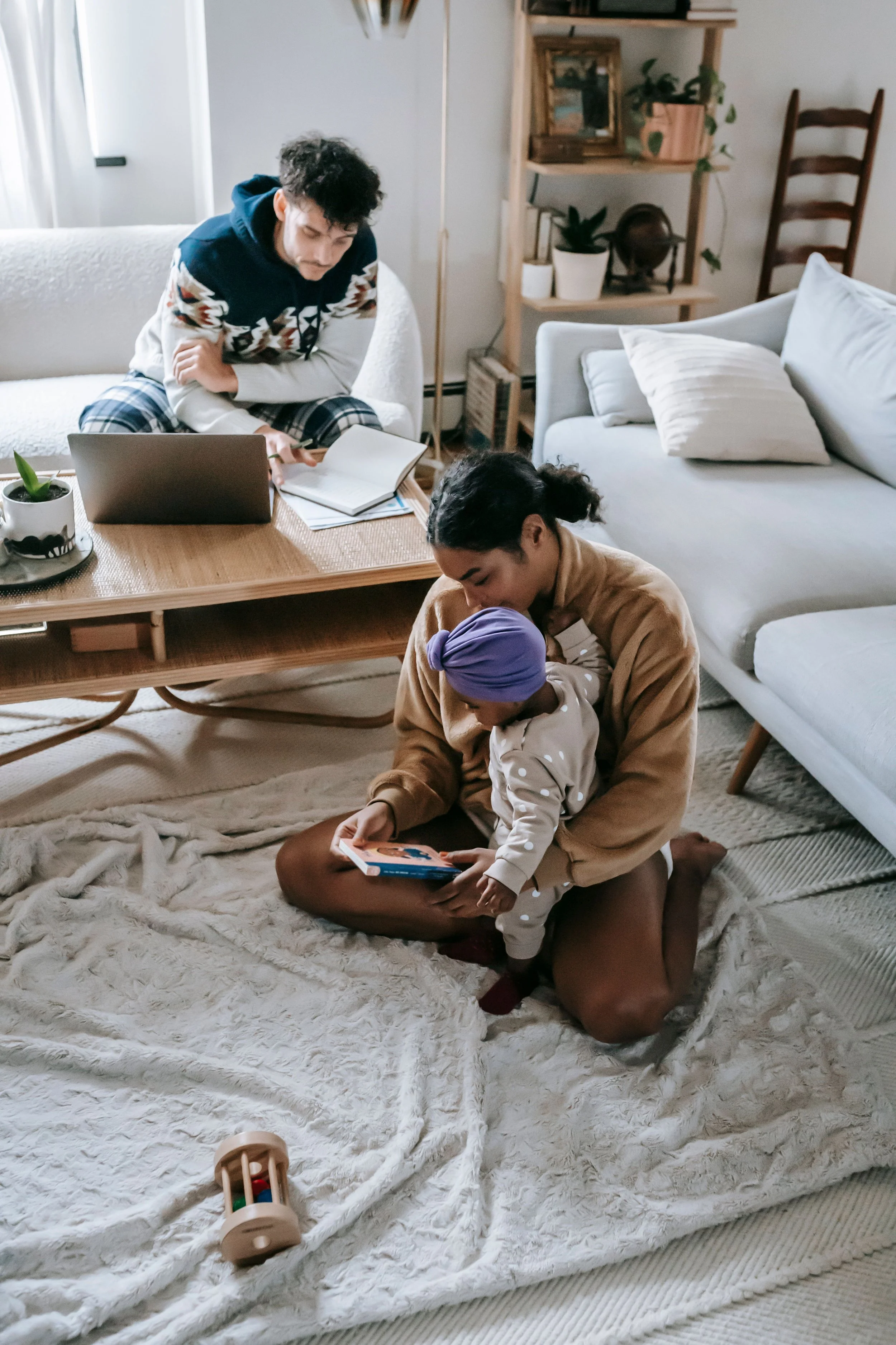 A Black mother sits on the floor with her child on her lap, reading a book together. In the background, the White father is sitting on a sofa behind a low table, looking down at papers. The room has a cozy, modern decor.