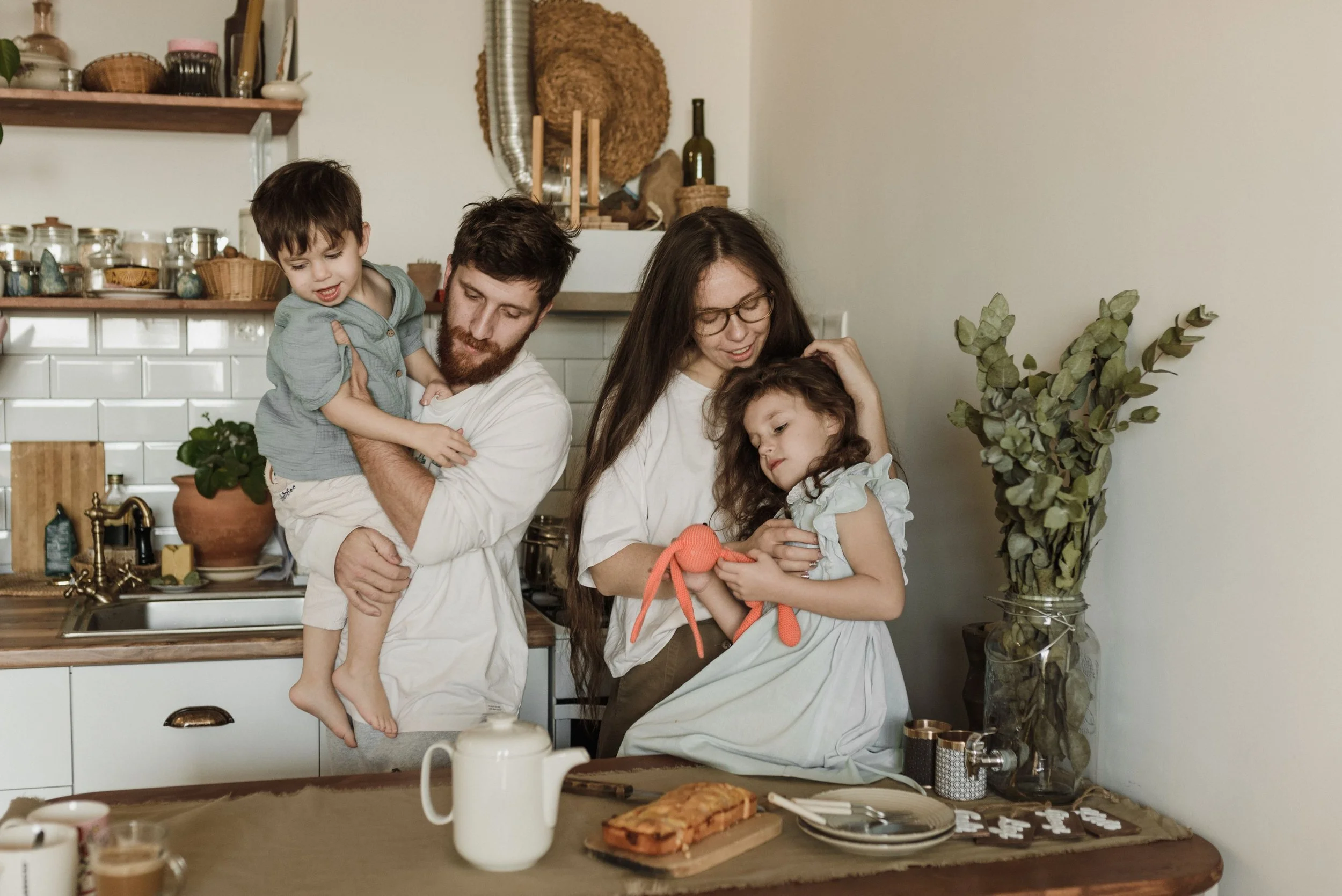 A family of four in a kitchen, with the white father holding a young boy and the light-skinned Latina mother holding a girl, all appearing to be engaged in a fun activity or game at the table. Shelves and plants are in the background.