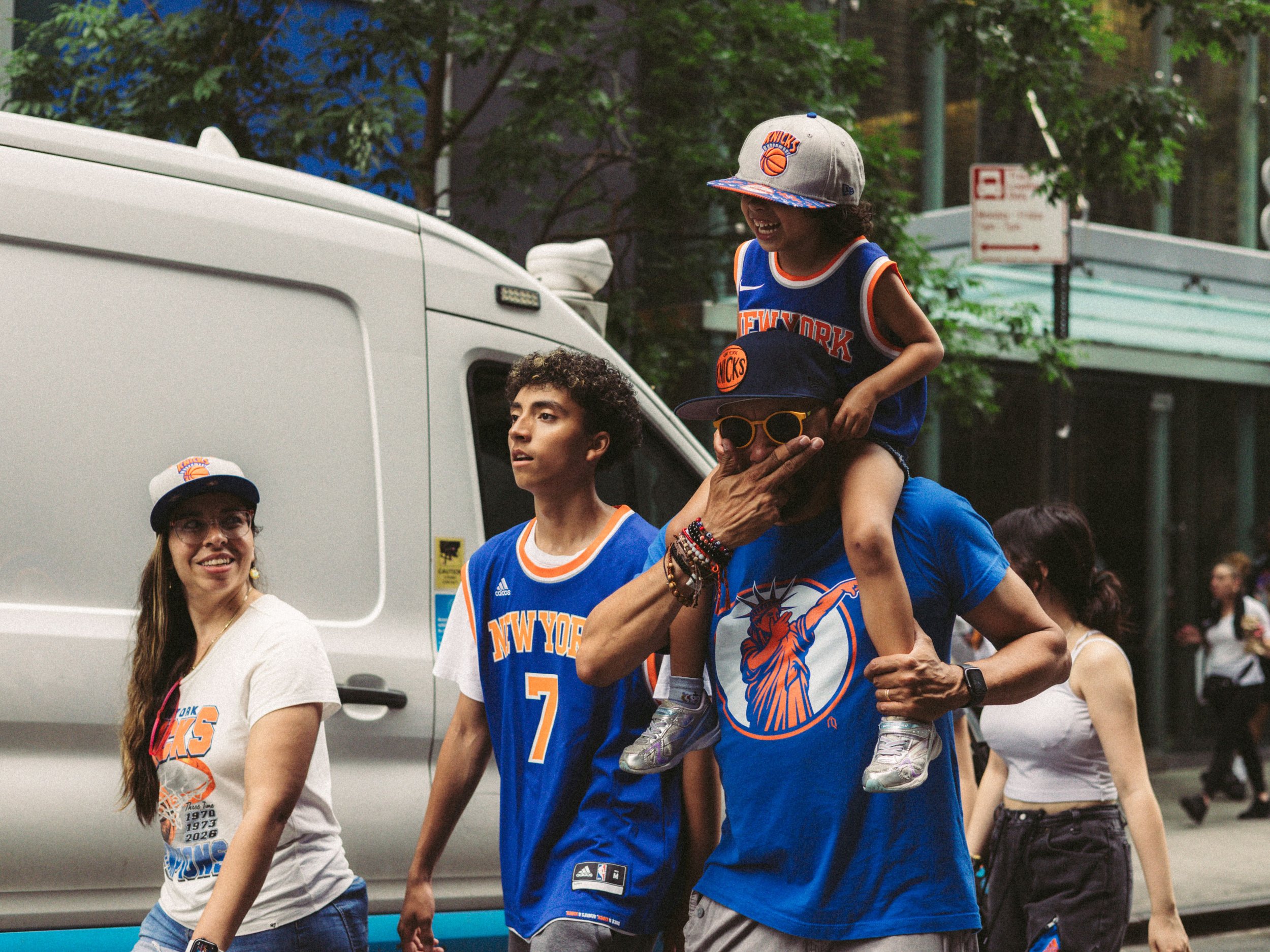A mixed race family walking and laughing together as if on the way to or from an event. The teenager, father, and the young child on his shoulders are wearing a New York Knicks shirts and caps. There are leafy trees and a white van in the background.