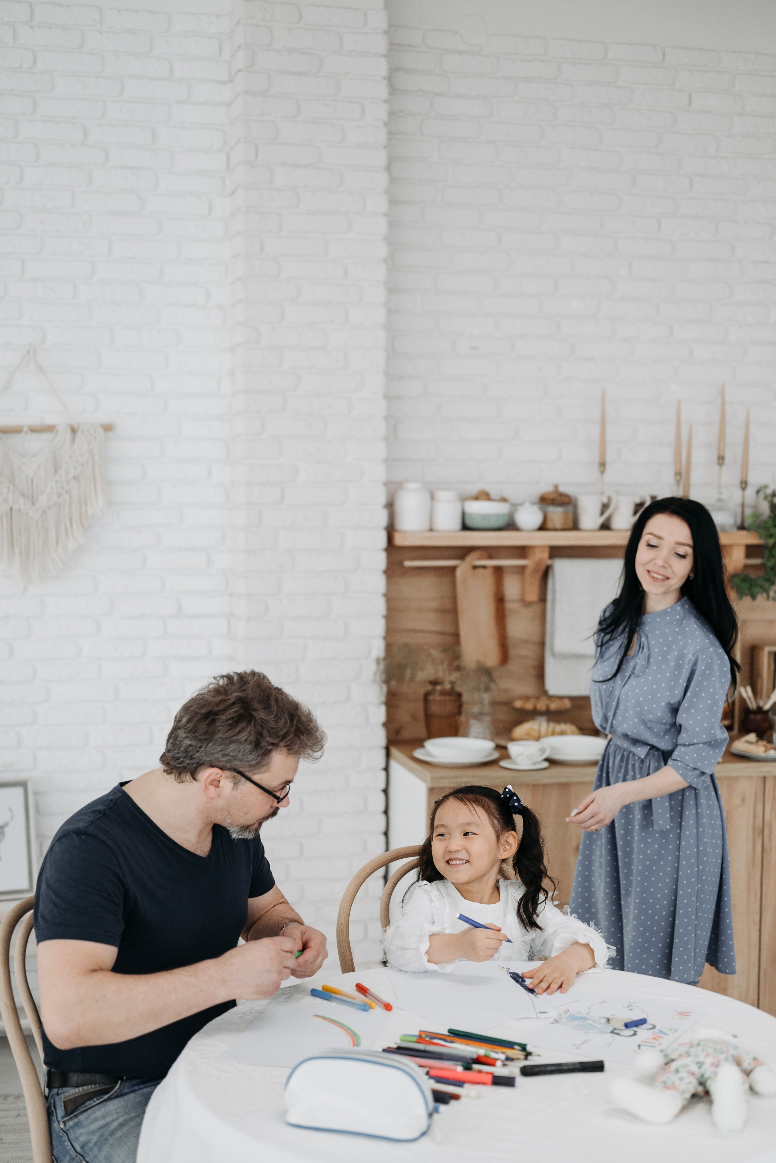 Family gathered around a table drawing and coloring with markers and colored pencils in a bright kitchen with white brick walls and wooden shelves.