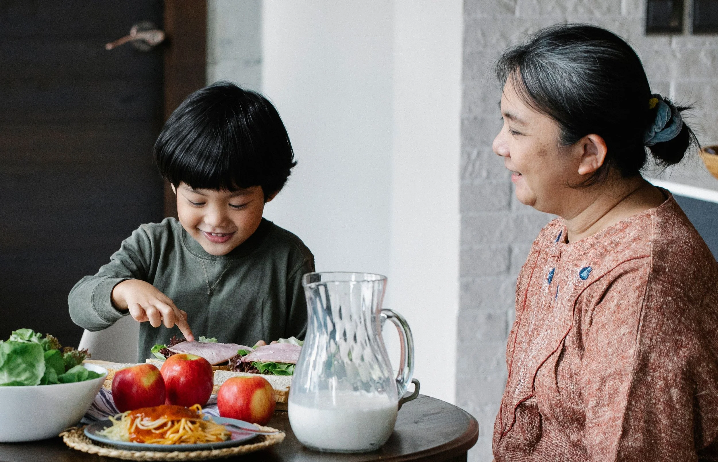 A young Asian boy with a bowl cut  sitting at a table with his grandmother, fresh vegetables and a pitcher of milk between them, smiling and sharing a moment together.