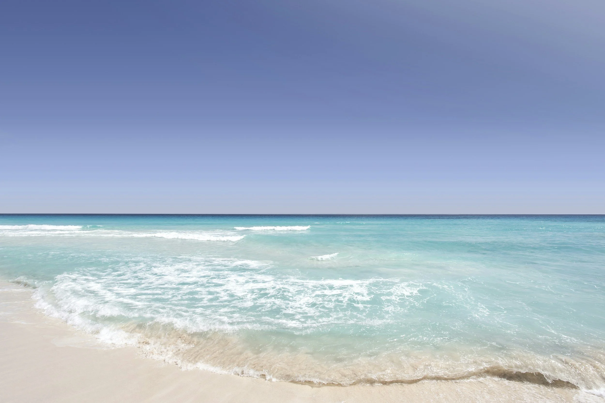 Calm ocean waves gently lapping a sandy beach with a clear blue sky overhead.