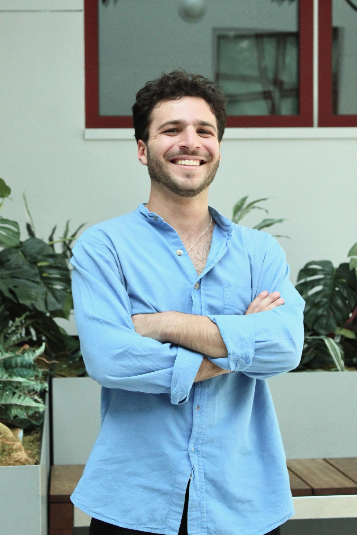 A young man with dark, curly hair and a beard smiling with arms crossed, wearing a light blue button-up shirt, standing in front of plants and a window in an indoor setting.