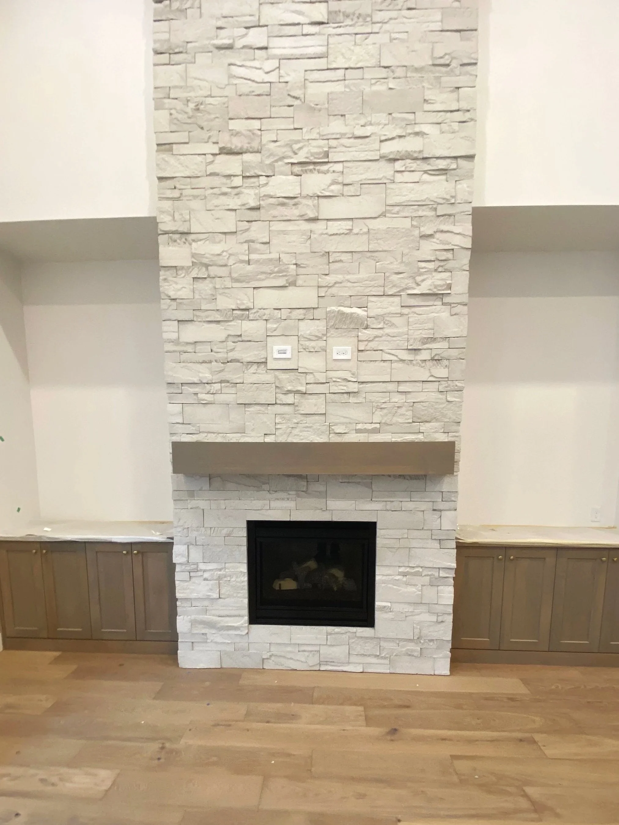 A modern white stone fireplace with a black firebox, flanked by built-in cabinetry and topped with a brown mantel. Wooden flooring is visible in front of the fireplace.