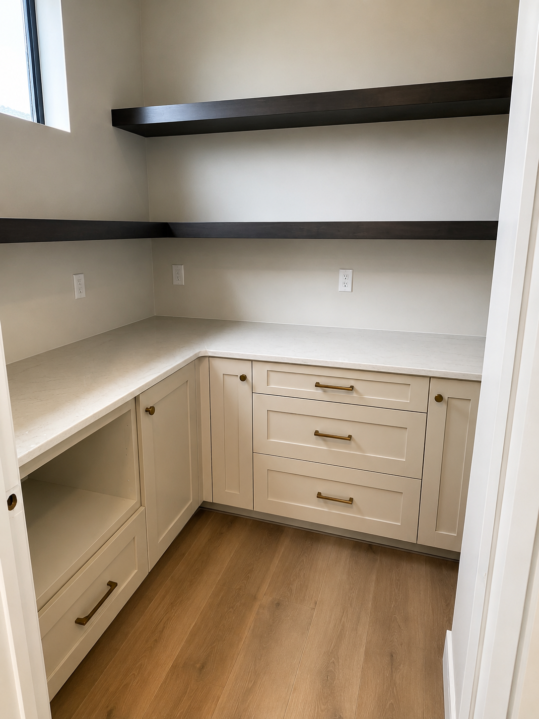 Empty kitchen corner with beige cabinets, gold handles, two open shelves, and three electrical outlets, with a window on the left and wood flooring.