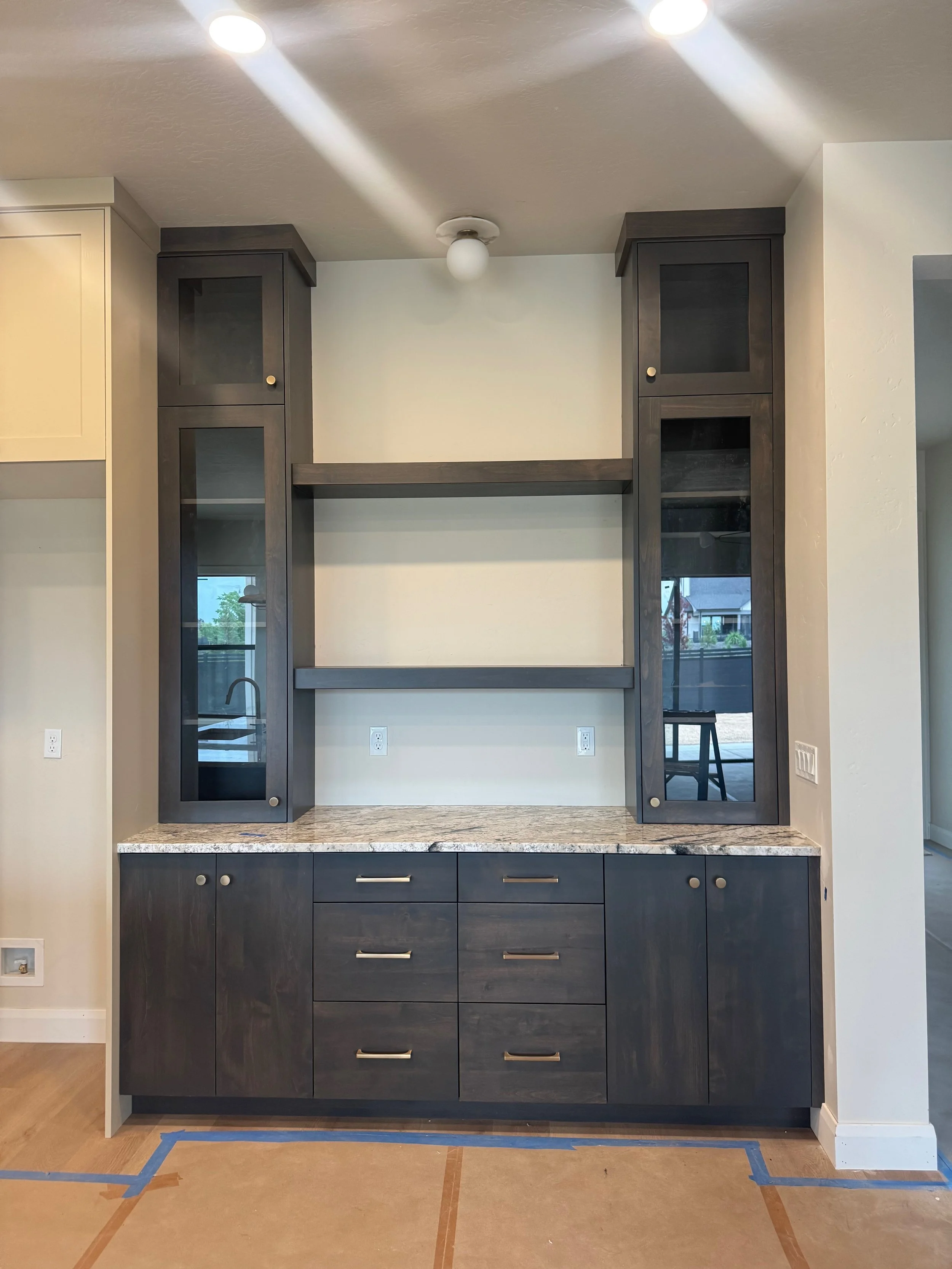 A built-in kitchen cabinet unit with dark wood finish, featuring glass-paneled upper cabinets, drawers, and lower cabinets on a tiled and papered floor. There is a granite countertop and electrical outlets on the wall.