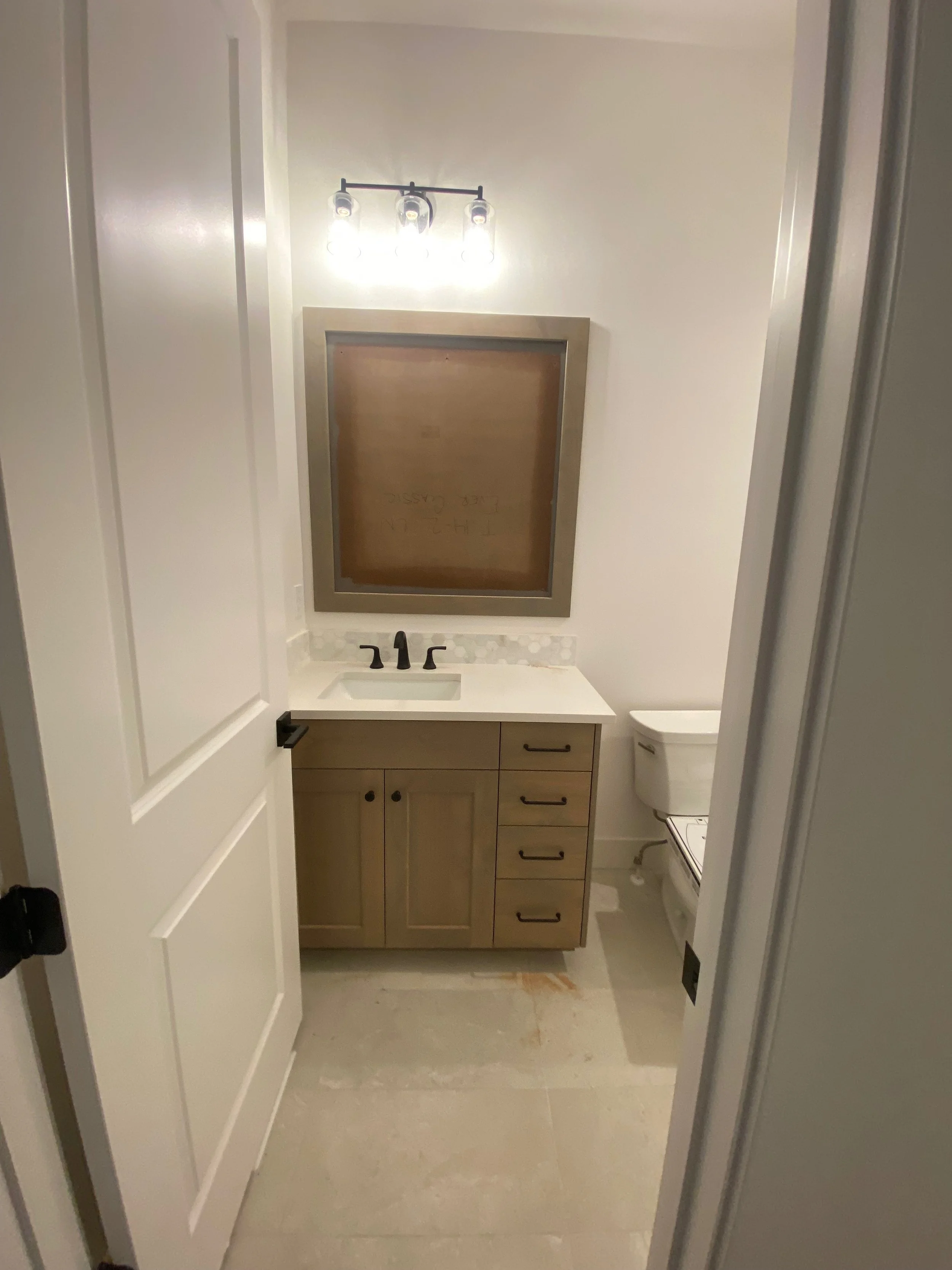 A bathroom with a white door open to reveal a wooden vanity with a white countertop and black hardware. There is a mirror above the sink and a modern light fixture with four exposed bulbs. A toilet is visible on the right side of the image, and the floor appears to be a light-colored tile or vinyl.
