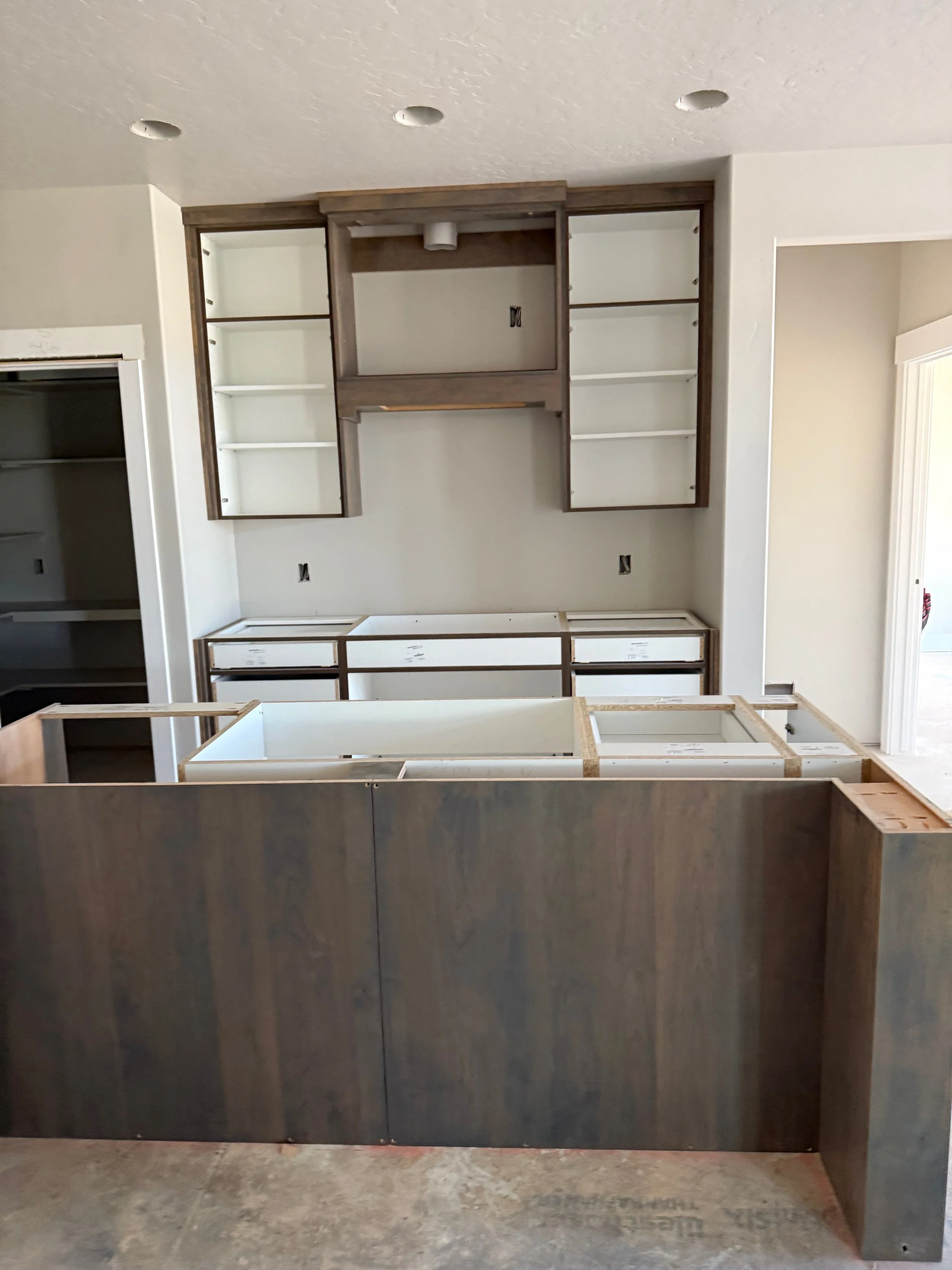 Kitchen cabinets in the process of installation, with open shelving and drawers yet to be assembled. The cabinets have a dark wood finish and are situated against a drywall wall with some electrical outlets and wiring visible.
