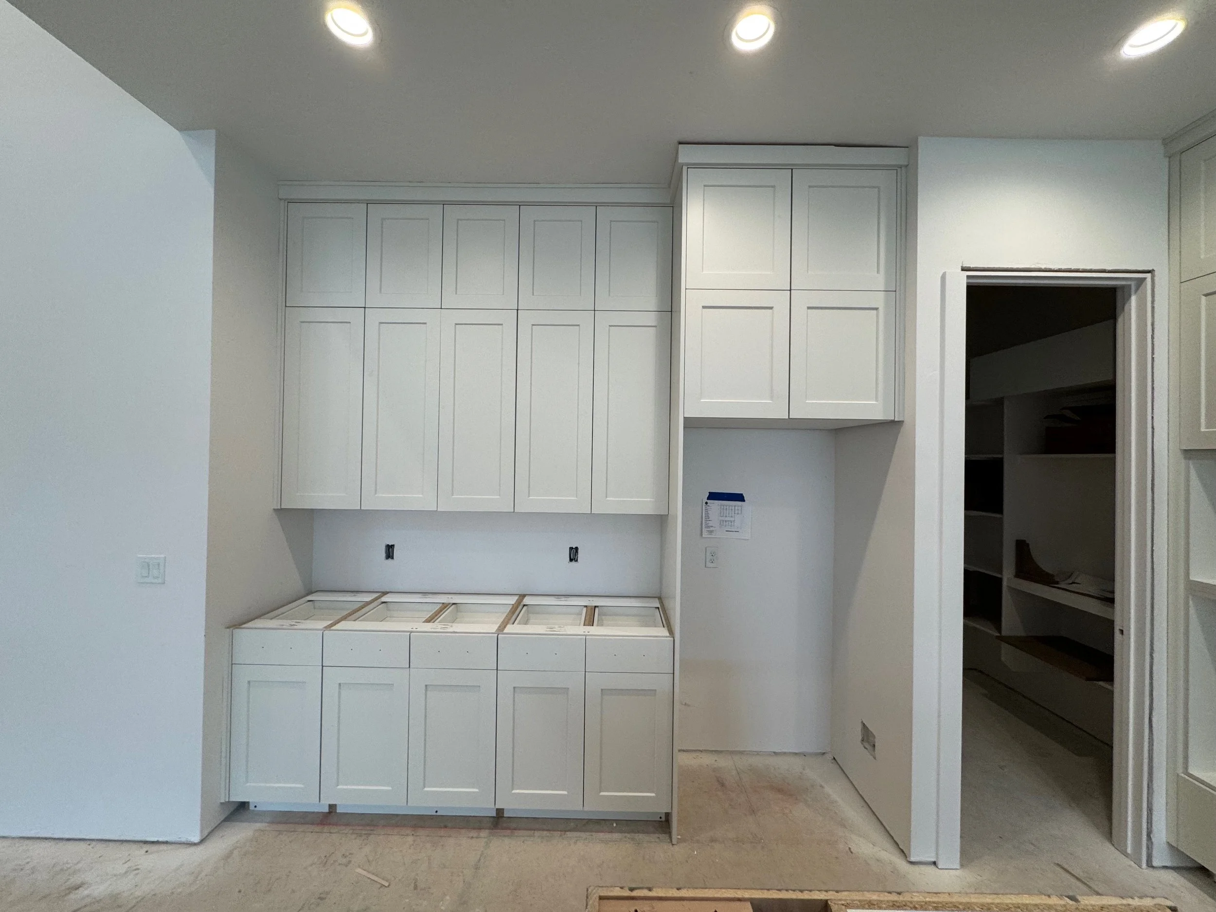 White kitchen cabinets are being installed, with some cabinets mounted on the wall and others on the floor, in an unfinished room with white walls and ceiling. Recessed lights are installed in the ceiling.