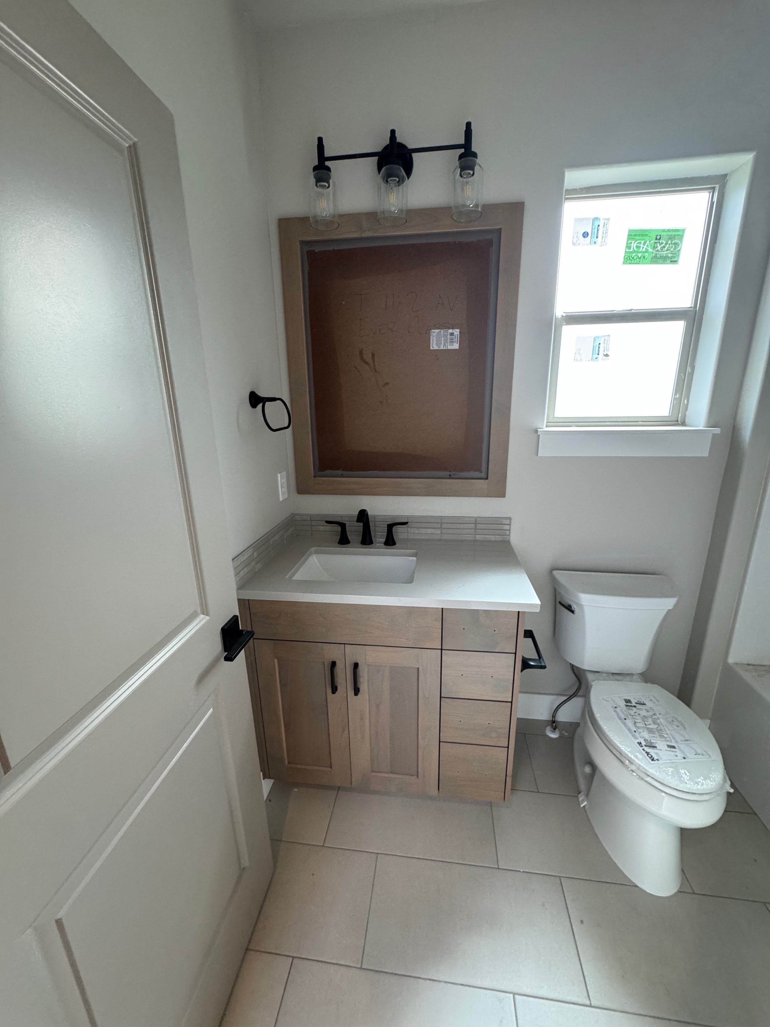Bathroom with a vanity mirror, light fixture with three bulbs, sink, toilet, and window. The mirror has a wooden frame, and the room has light-colored tiles and walls.